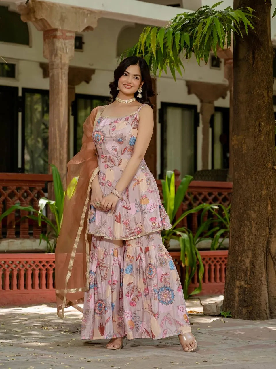 Woman in a floral dress standing in an outdoor setting with plants and architectural elements.