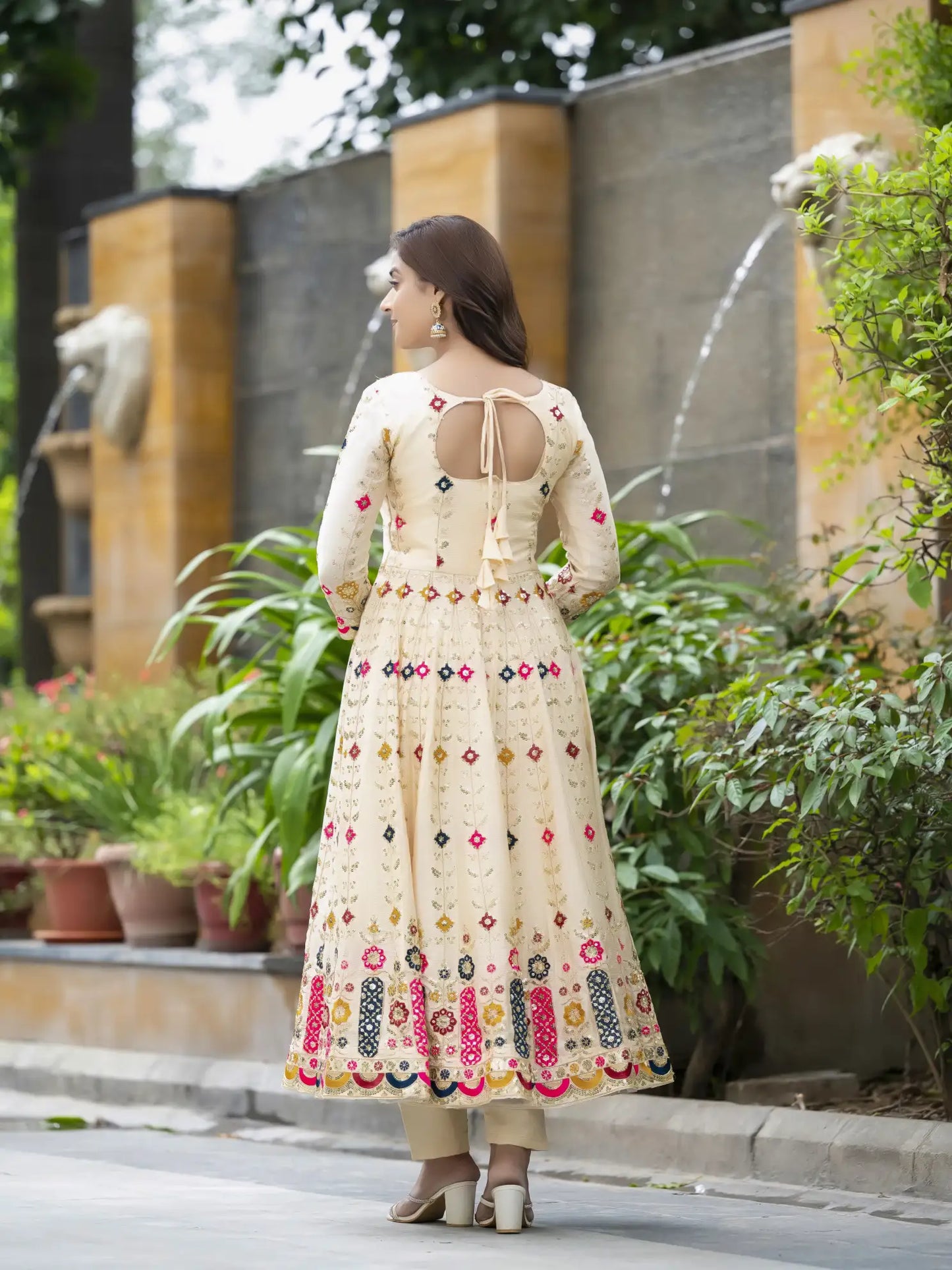 Woman in a cream-colored dress with floral patterns walking outdoors near a water fountain.