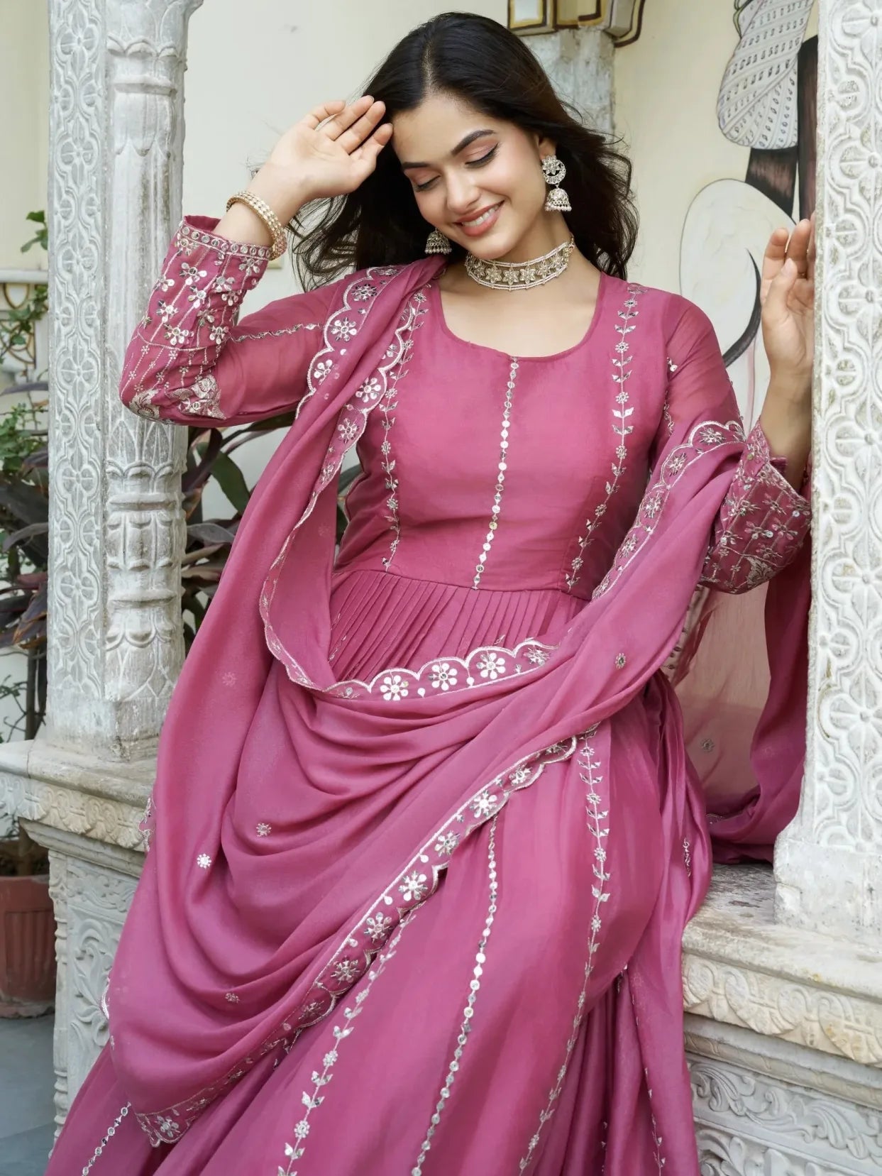 Woman in a pink traditional outfit with intricate designs, standing against an ornate white background.
