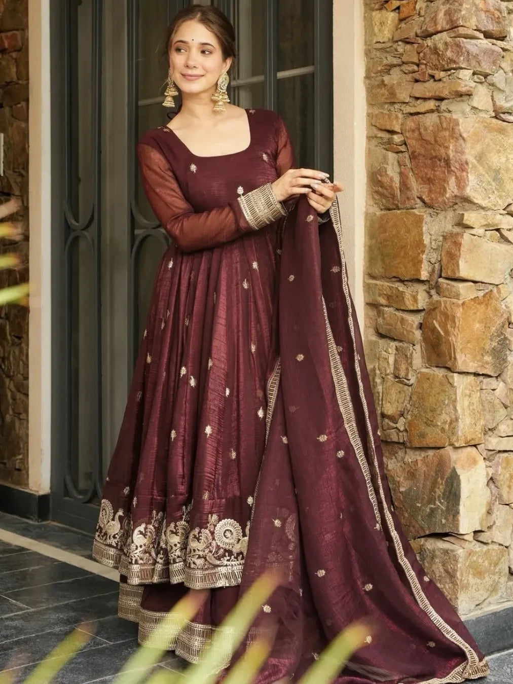 Woman in a maroon traditional outfit standing in front of a stone wall.
