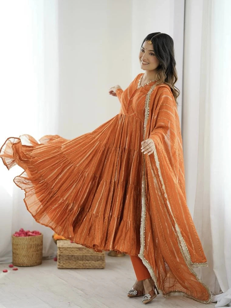 Woman in an orange traditional outfit with a flowing design against a white curtain background.