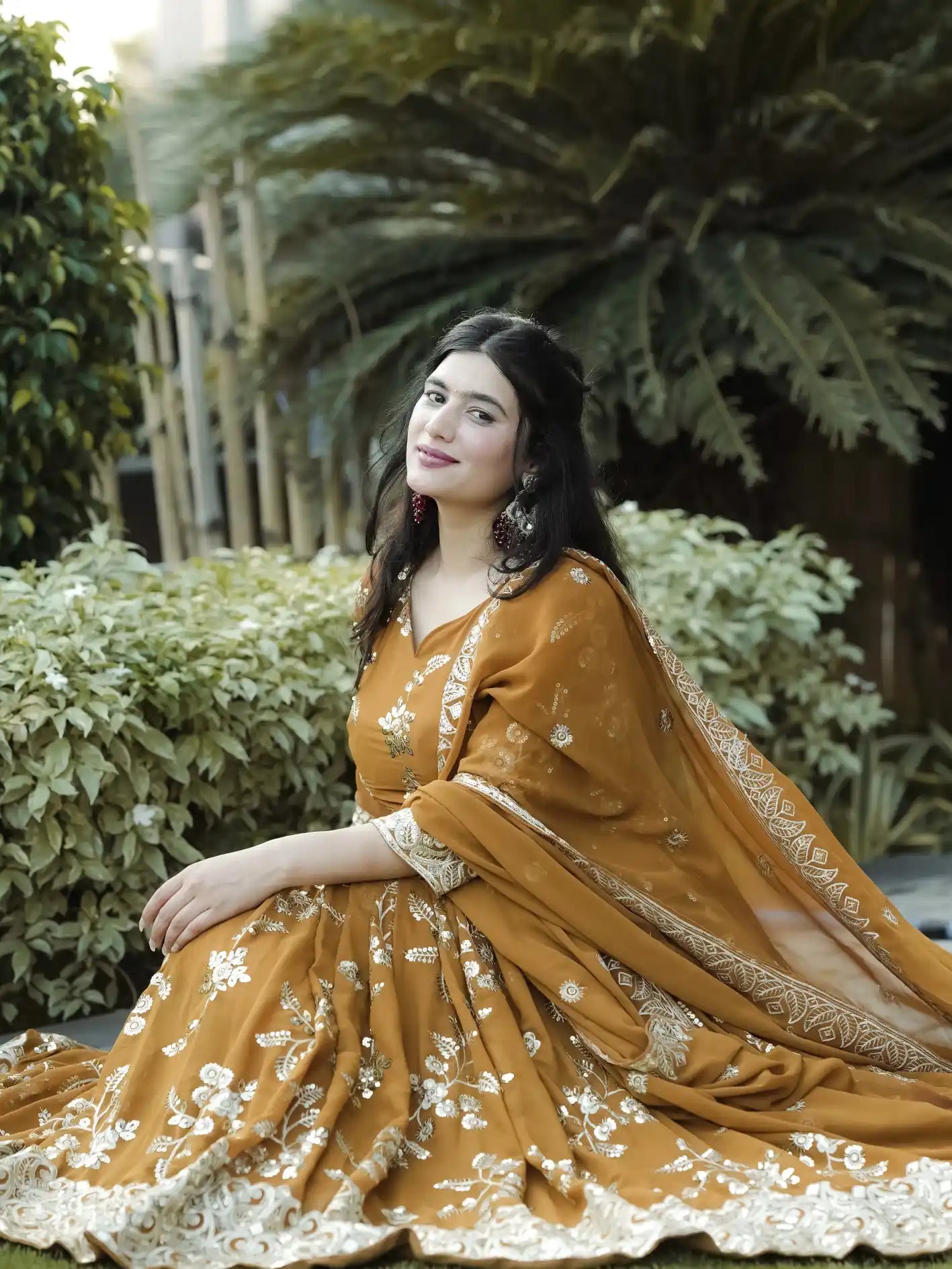 Woman in a mustard yellow traditional outfit with floral patterns sitting outdoors among greenery.