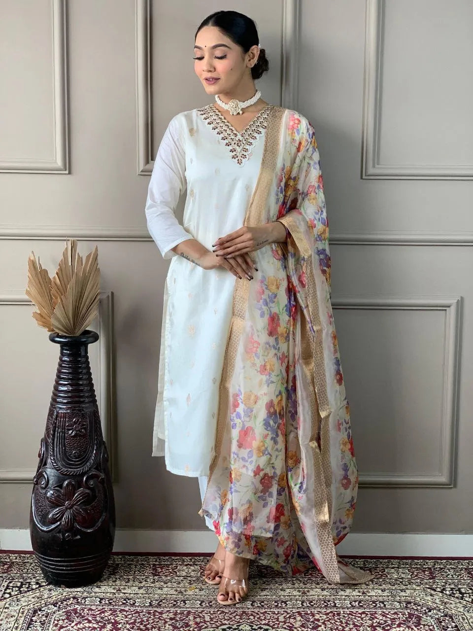 Woman in traditional outfit with floral dupatta standing against a neutral wall.