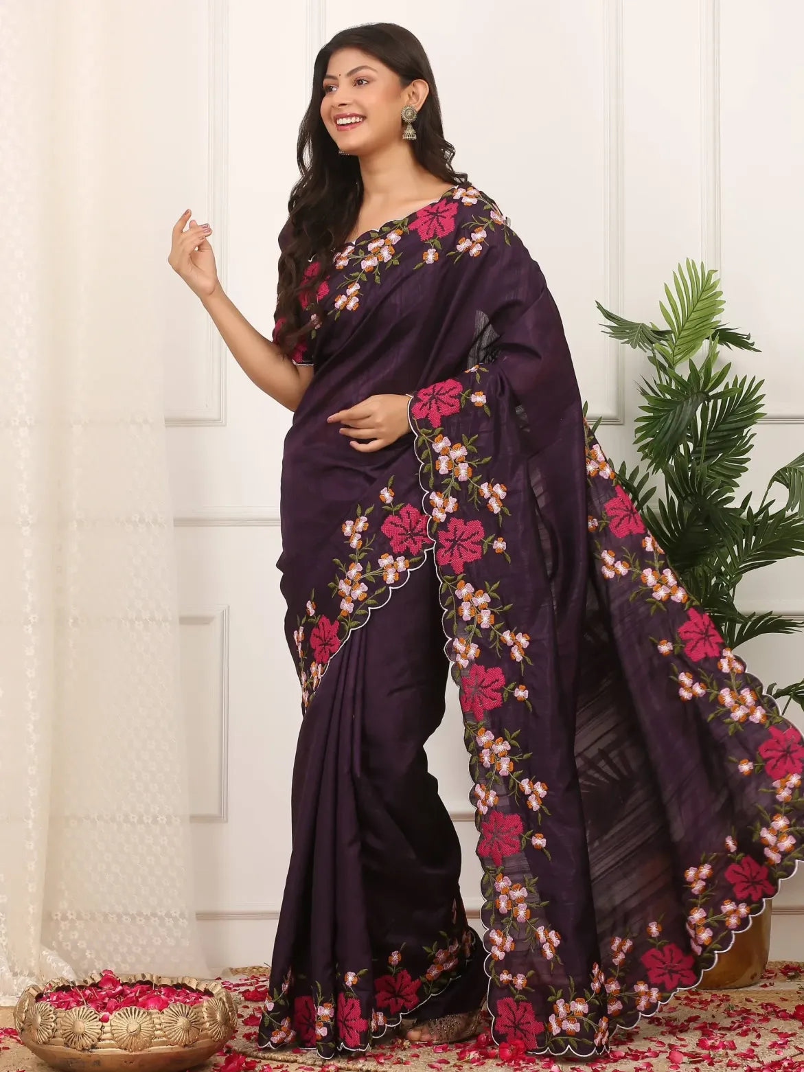 Woman wearing a dark purple saree with floral embroidery against a white curtain background.