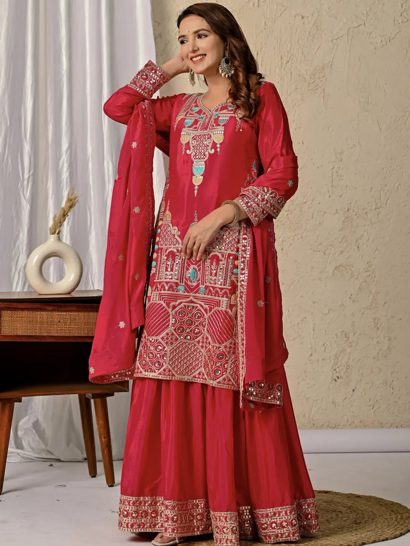 Woman wearing a red embroidered traditional outfit with a beige wall and decorative items in the background