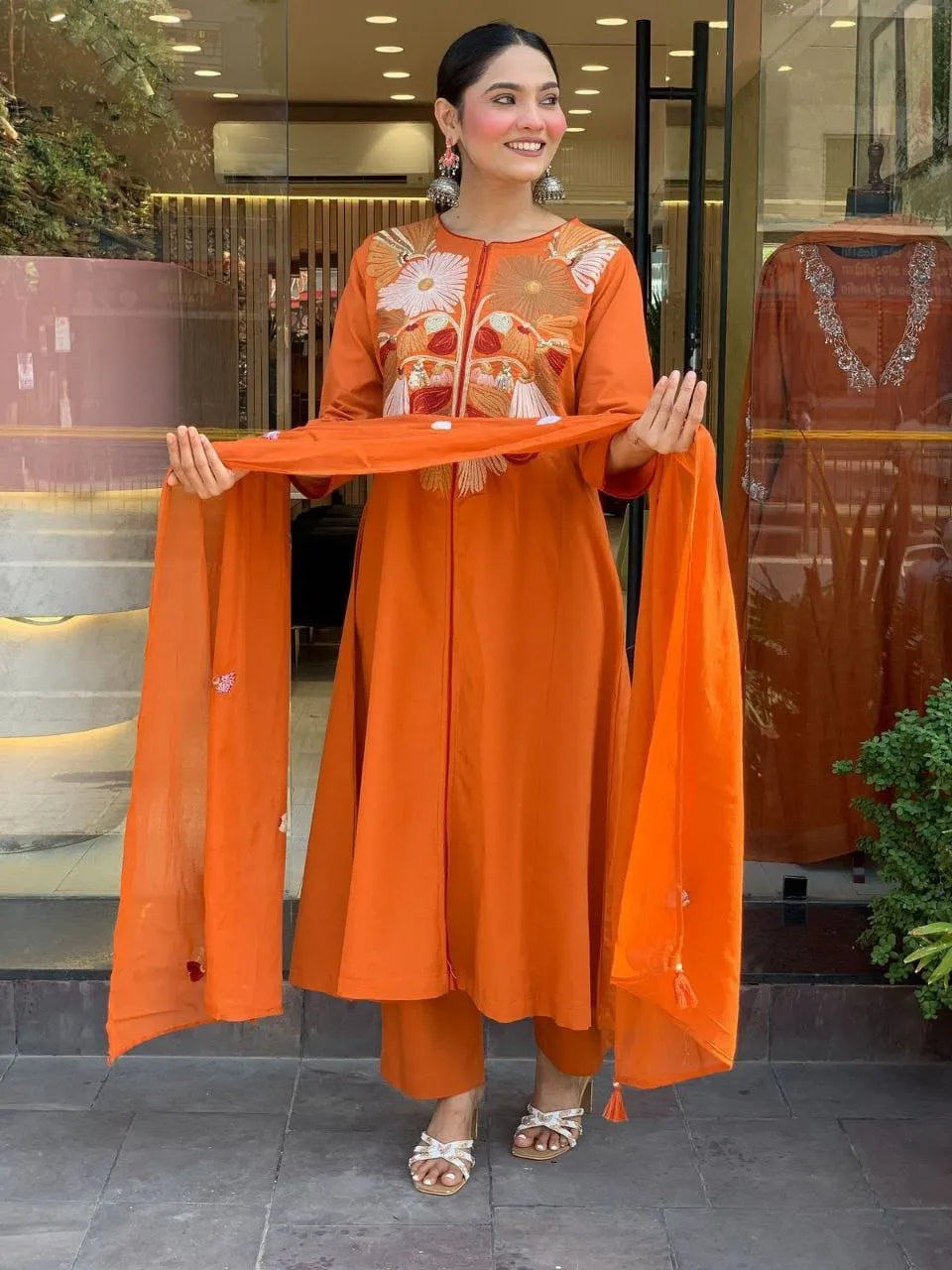 Woman in an orange traditional outfit holding a matching dupatta, standing in front of a glass door.