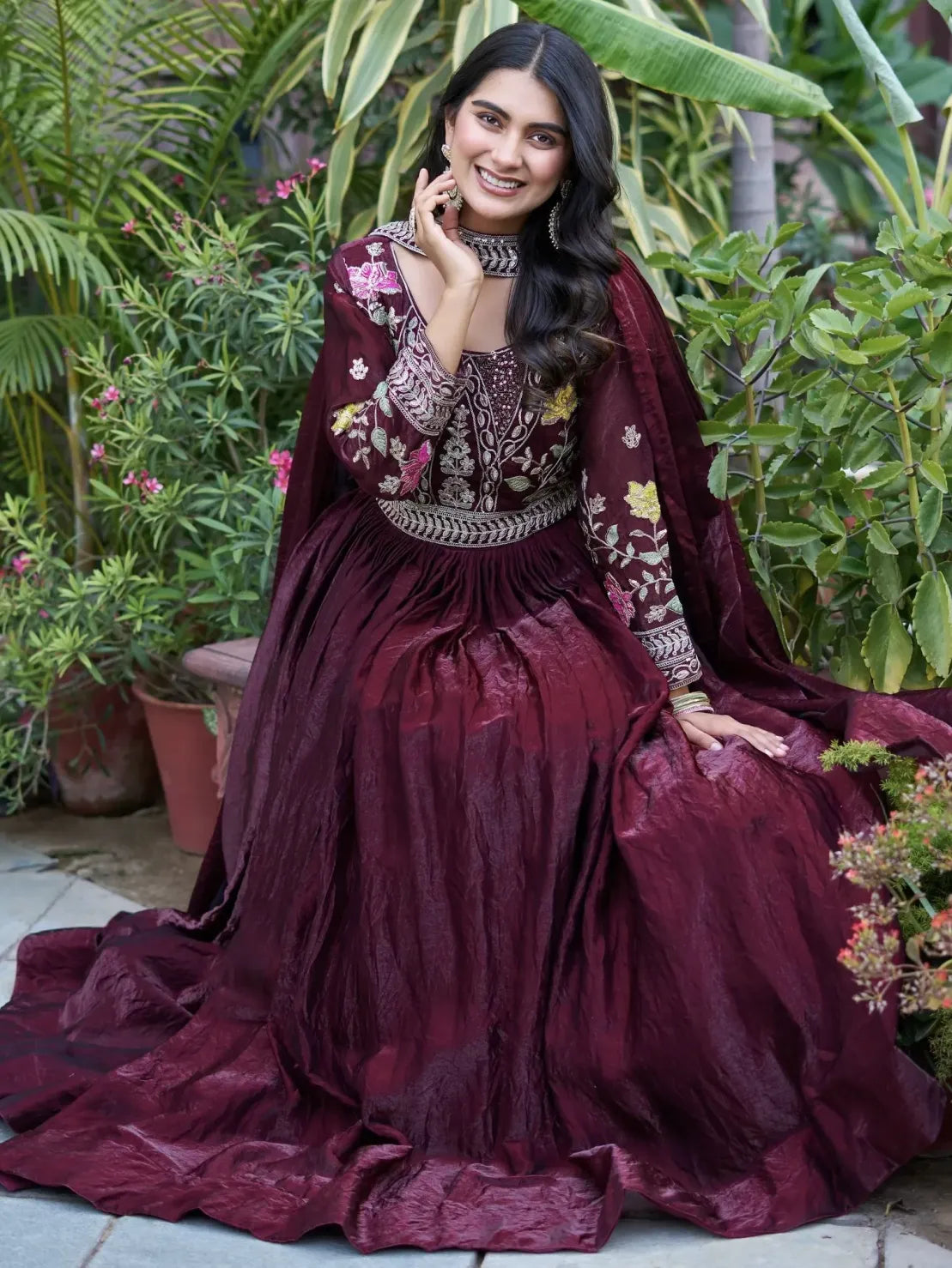 Woman in a burgundy embroidered traditional outfit sitting among plants.