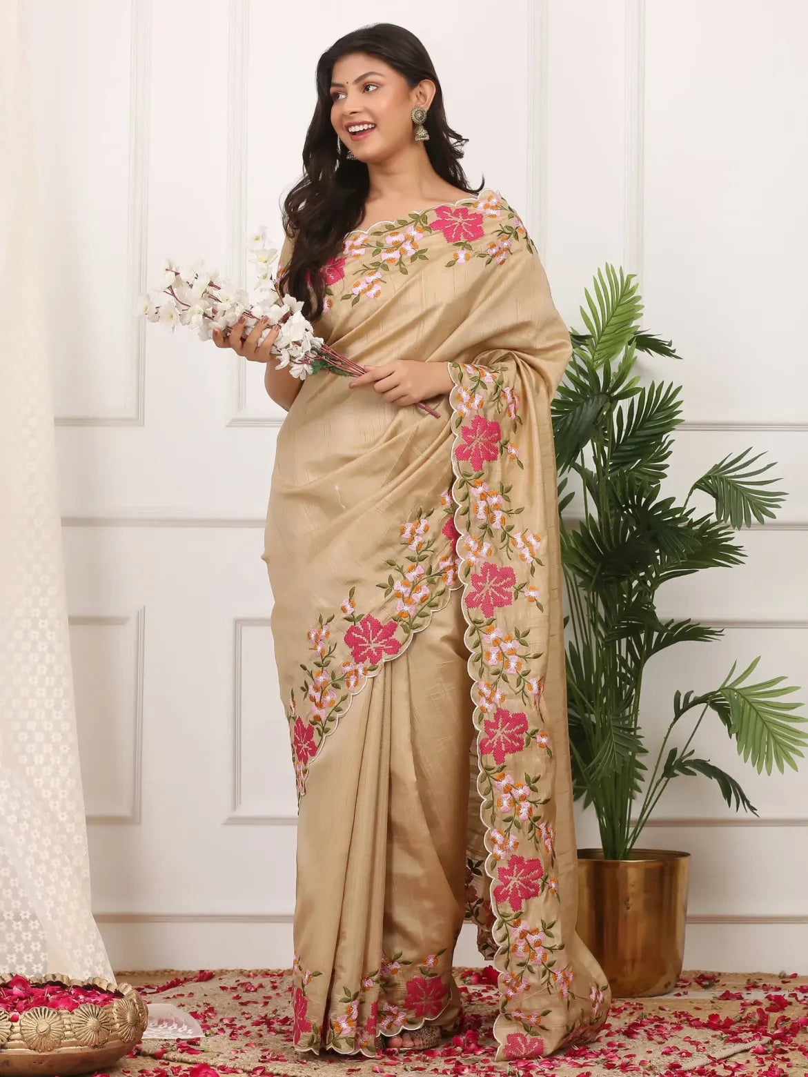 Woman wearing a beige saree with floral embroidery, holding flowers indoors.