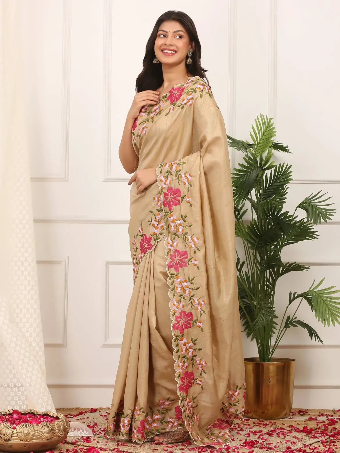 Woman wearing a beige saree with floral embroidery in a room with a plant and decorative rug.