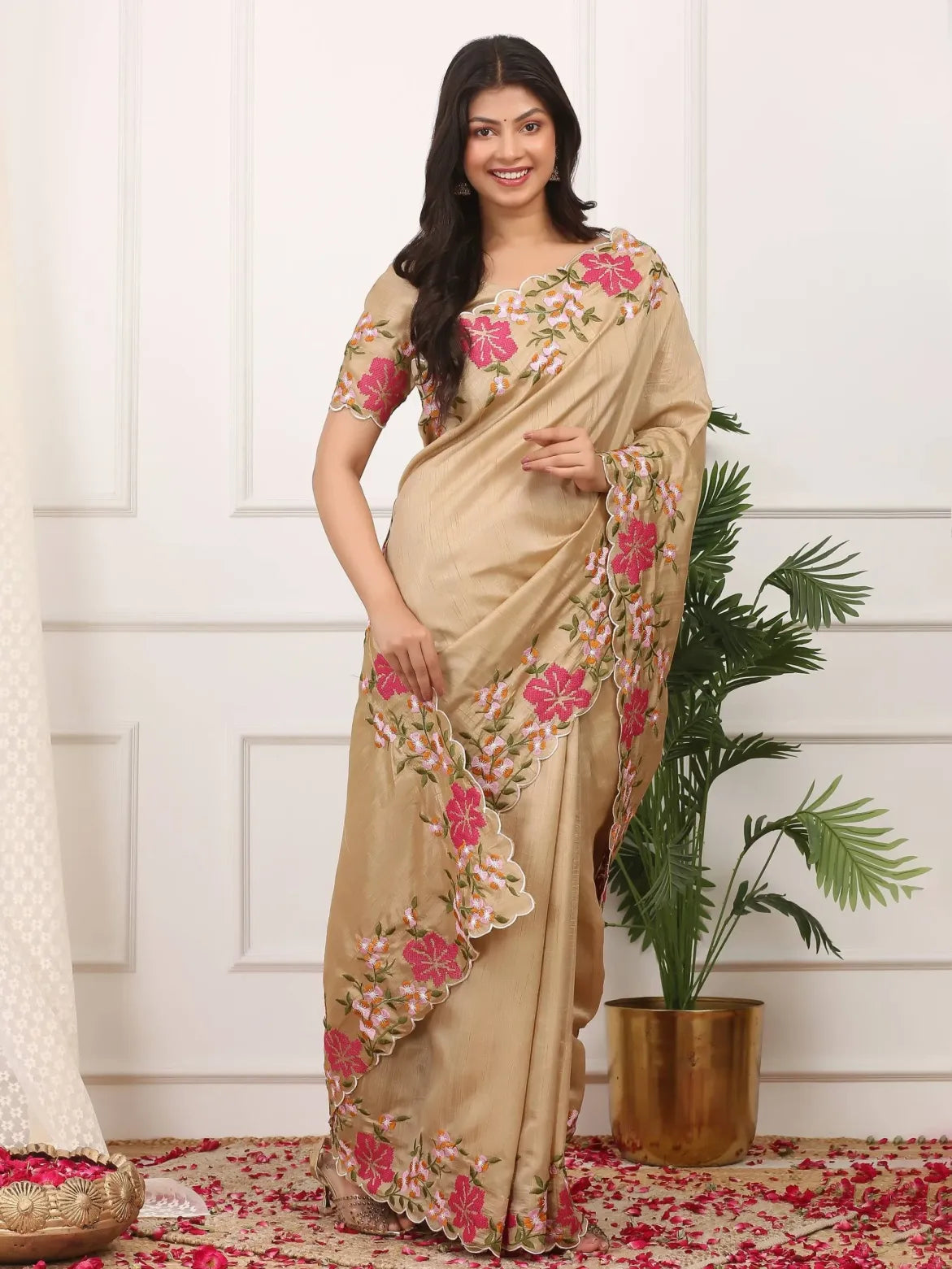 Woman wearing a beige saree with floral patterns in a room with a plant and rug.