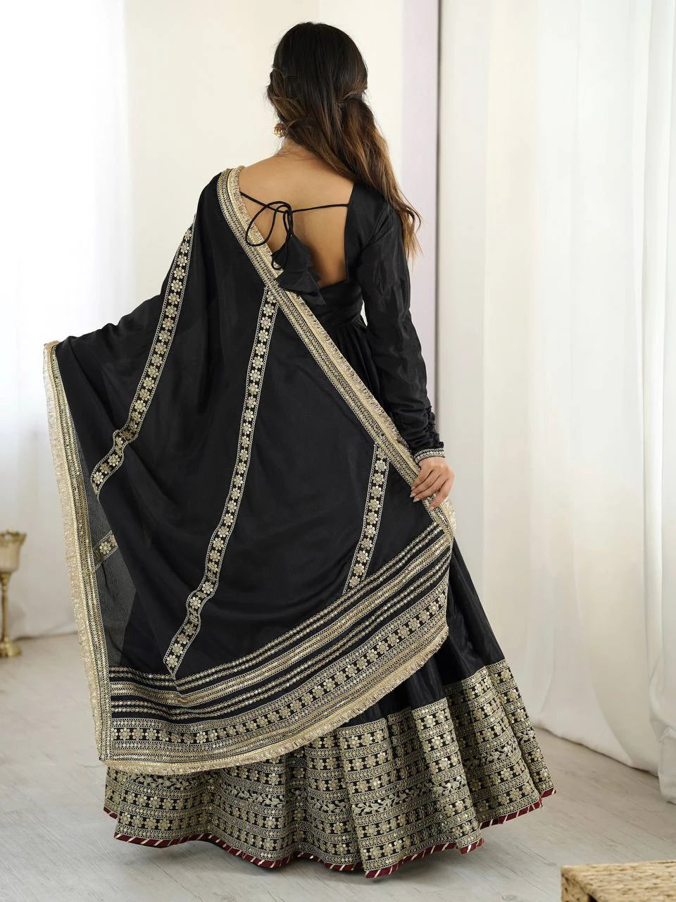 Woman wearing a black saree with intricate patterns, standing against a white curtain.