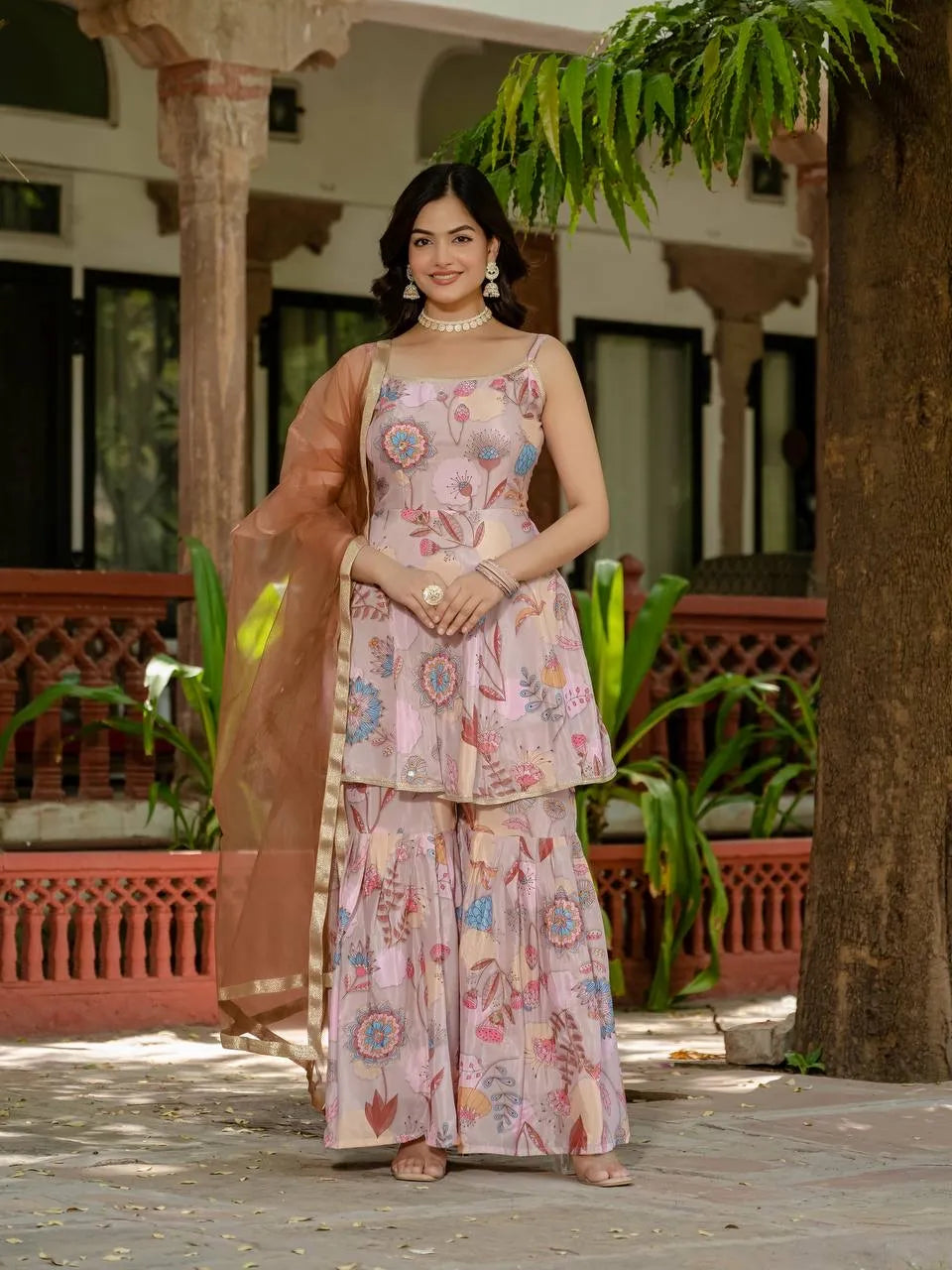 Woman in a floral outfit standing outdoors with plants and architectural elements in the background