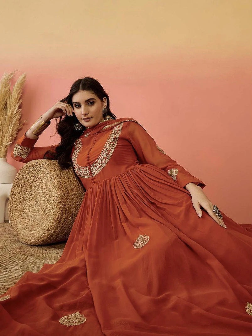 Woman in a rust-colored traditional outfit with gold embroidery against a peach wall.