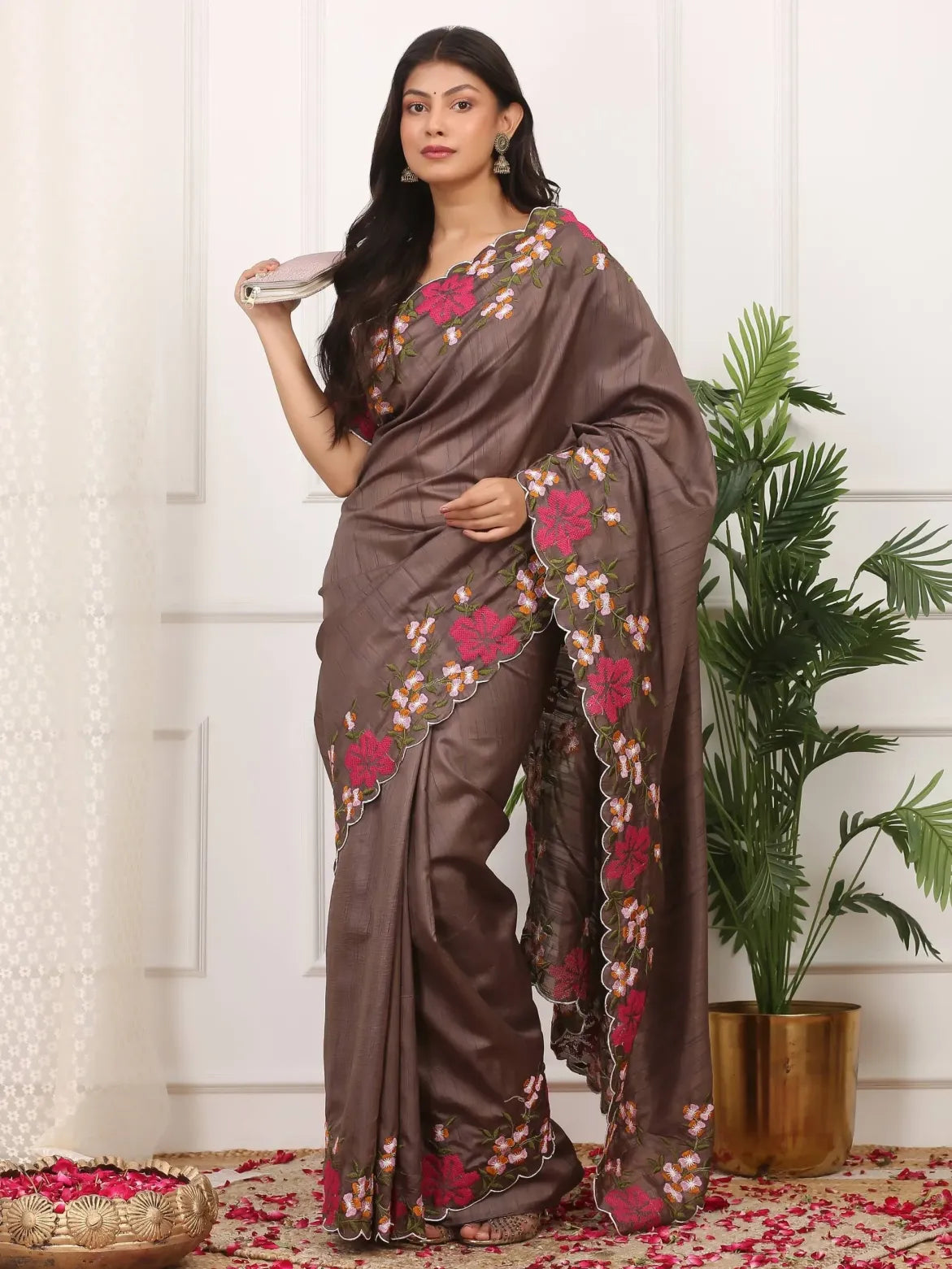 Woman wearing a brown saree with floral patterns, standing against a white wall with a plant in the background.