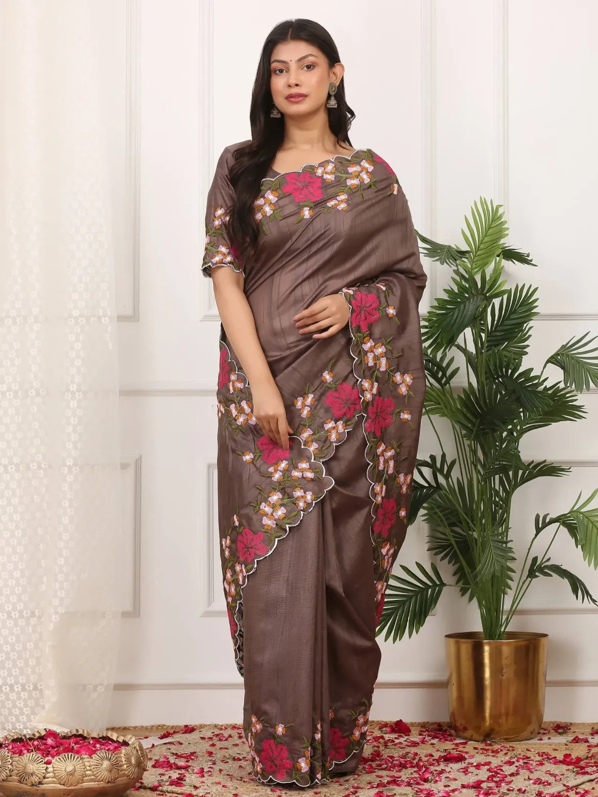 Woman wearing a brown saree with floral patterns standing indoors next to a plant.