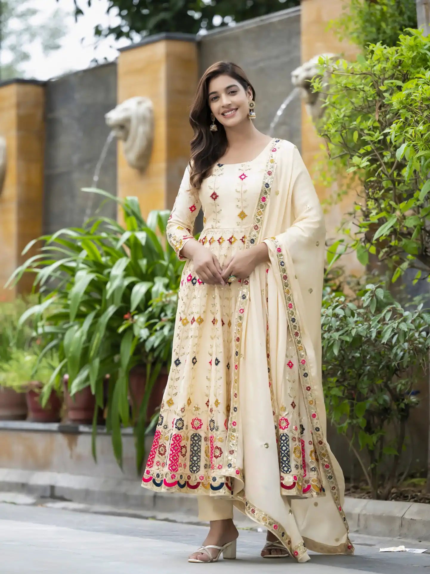 Woman in a cream-colored embroidered traditional outfit standing outdoors with greenery in the background