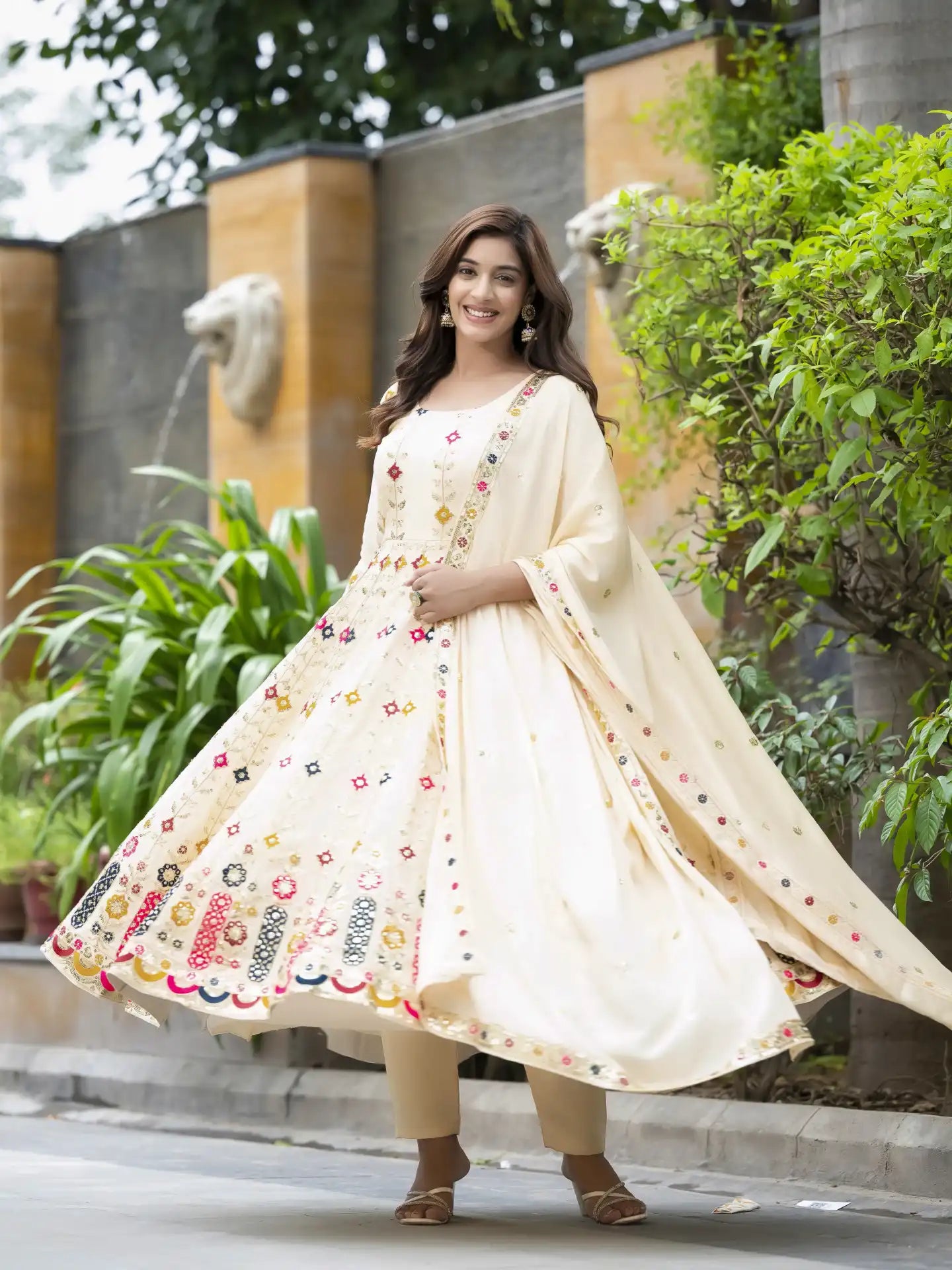 Woman in a cream-colored embroidered anarkali suit standing outdoors with greenery in the background