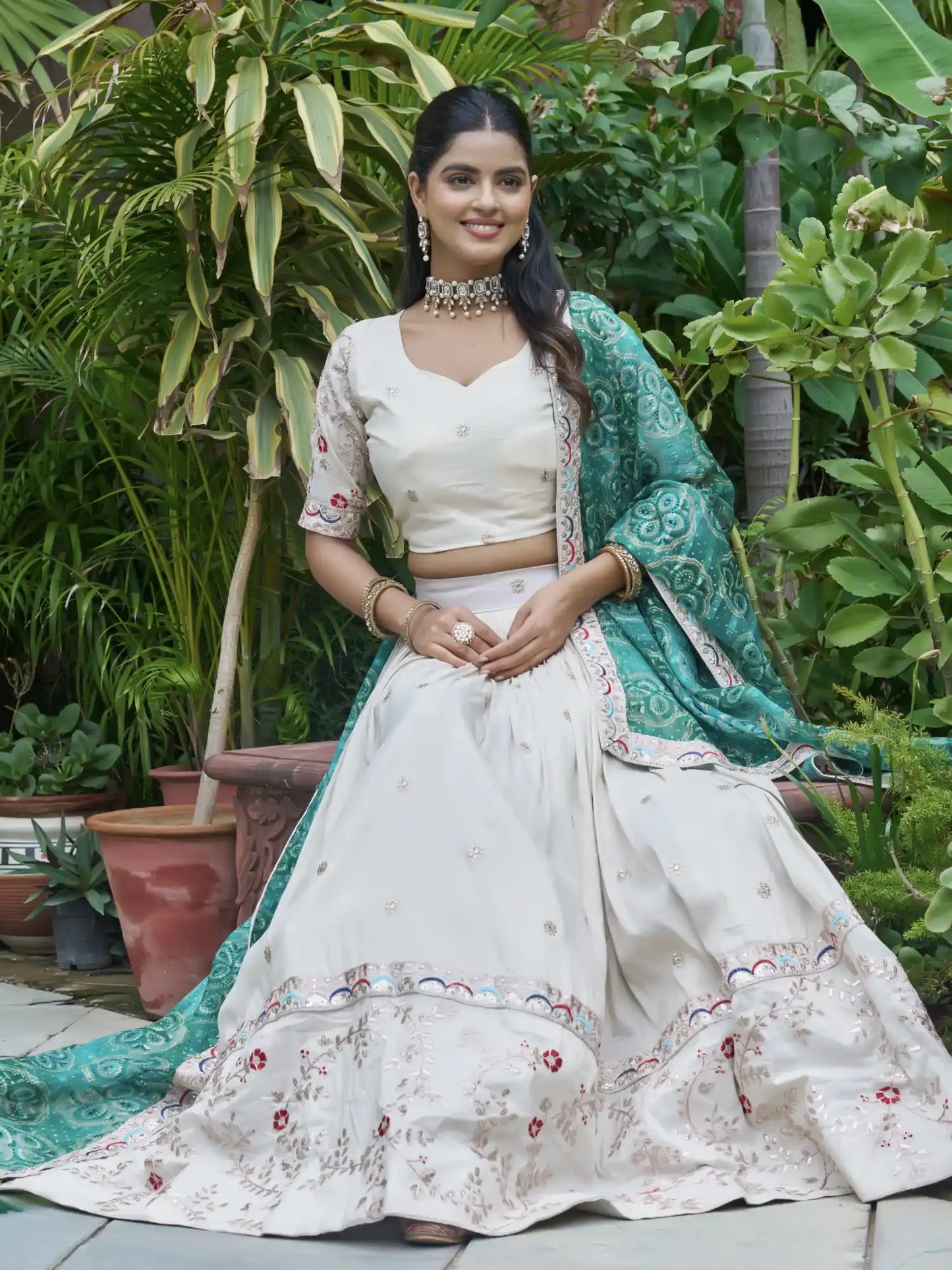 Woman in a white and green traditional outfit sitting outdoors with plants in the background