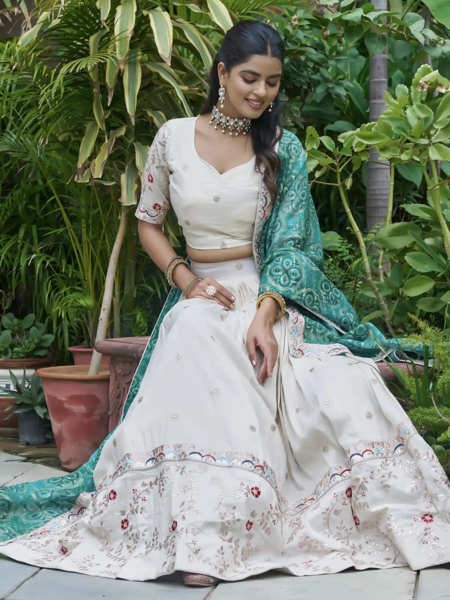 Woman in a white saree with floral patterns sitting outdoors among greenery