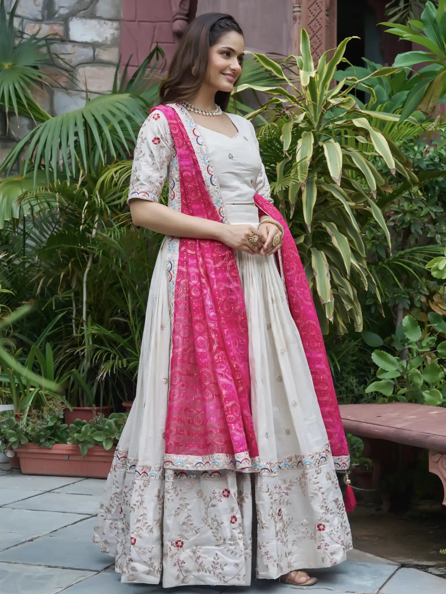 Woman in a white and pink traditional outfit standing outdoors with greenery.