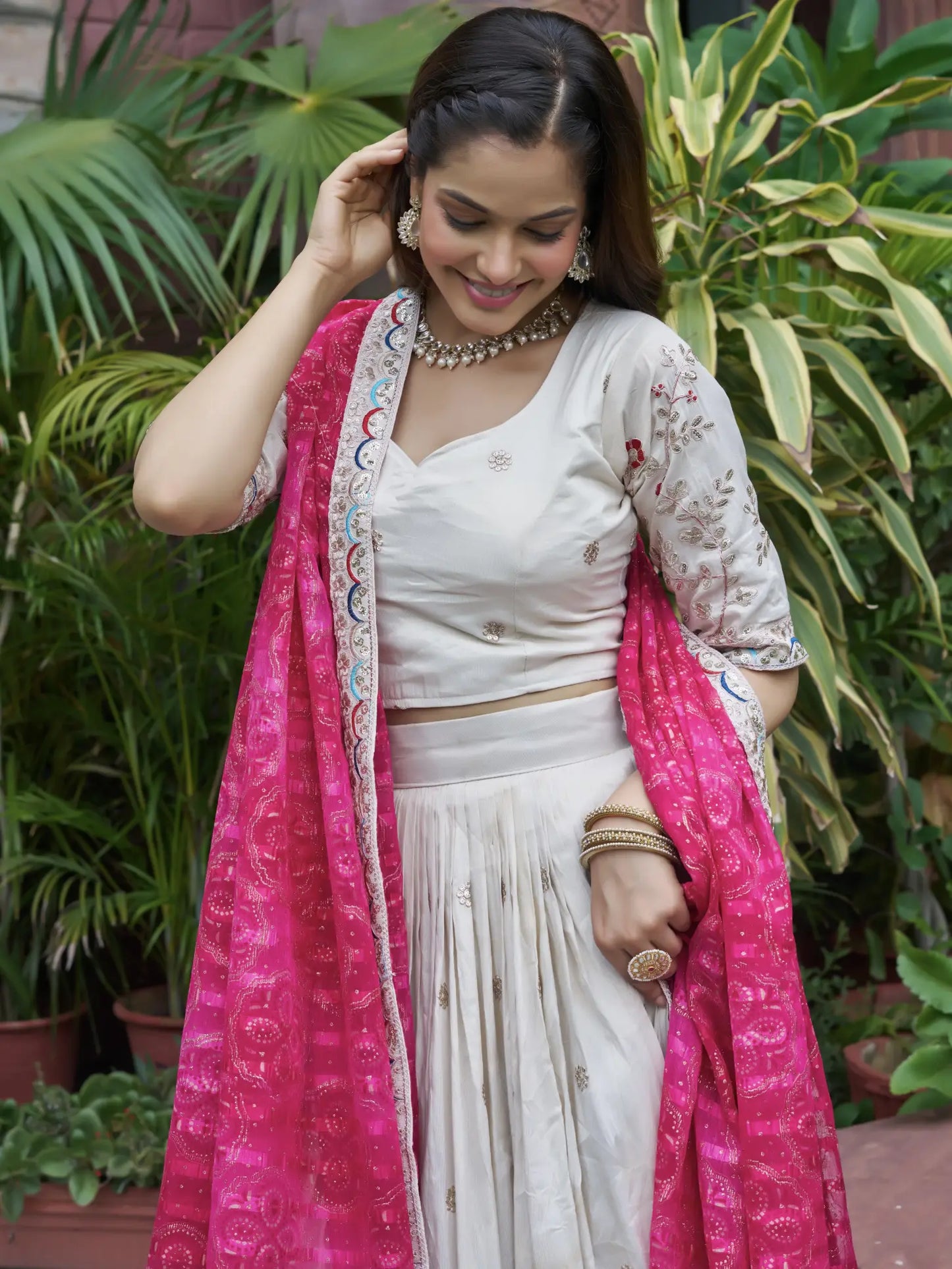 Woman in a white traditional outfit with a pink dupatta standing among plants.