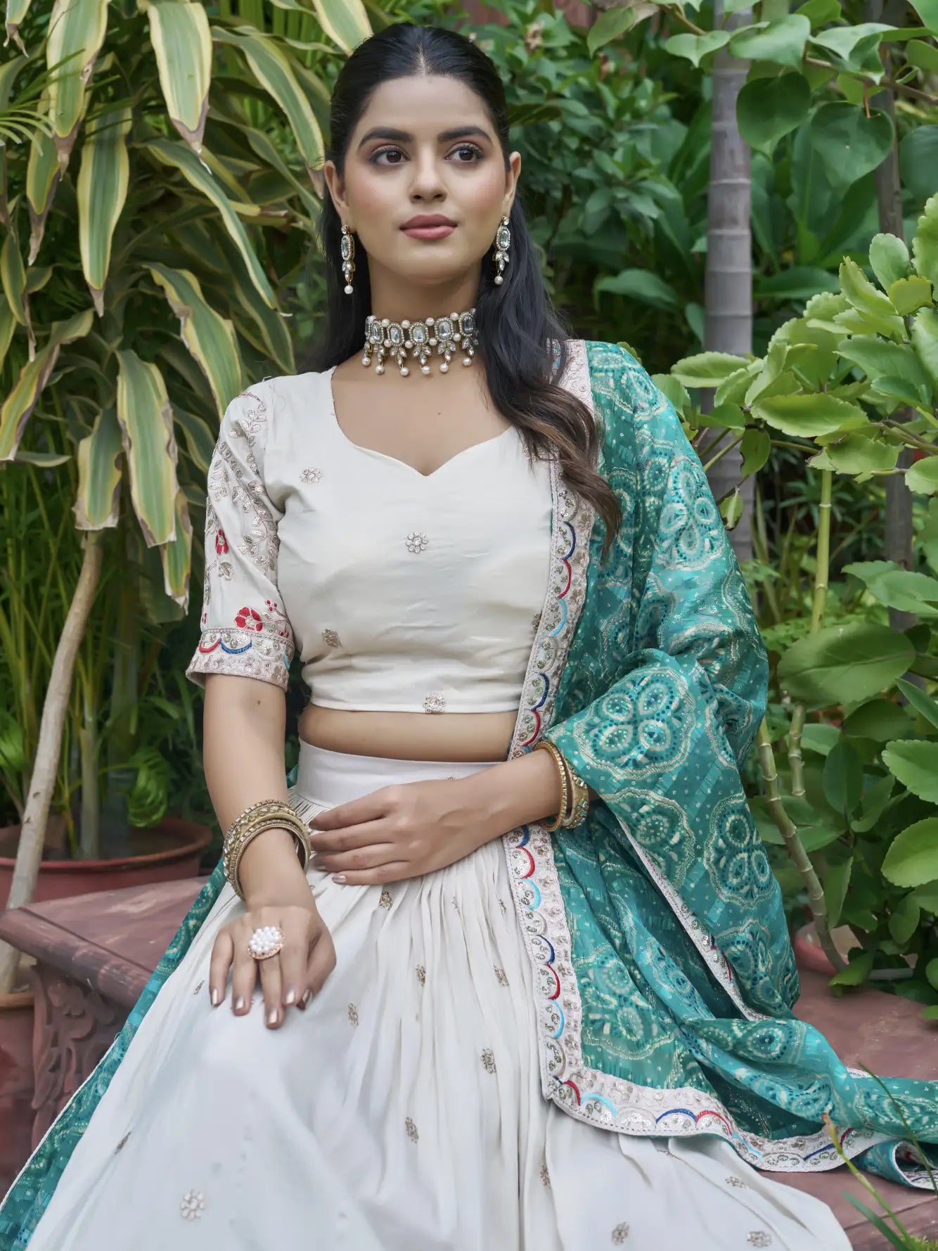 Woman in a white and green traditional outfit with jewelry, sitting outdoors.