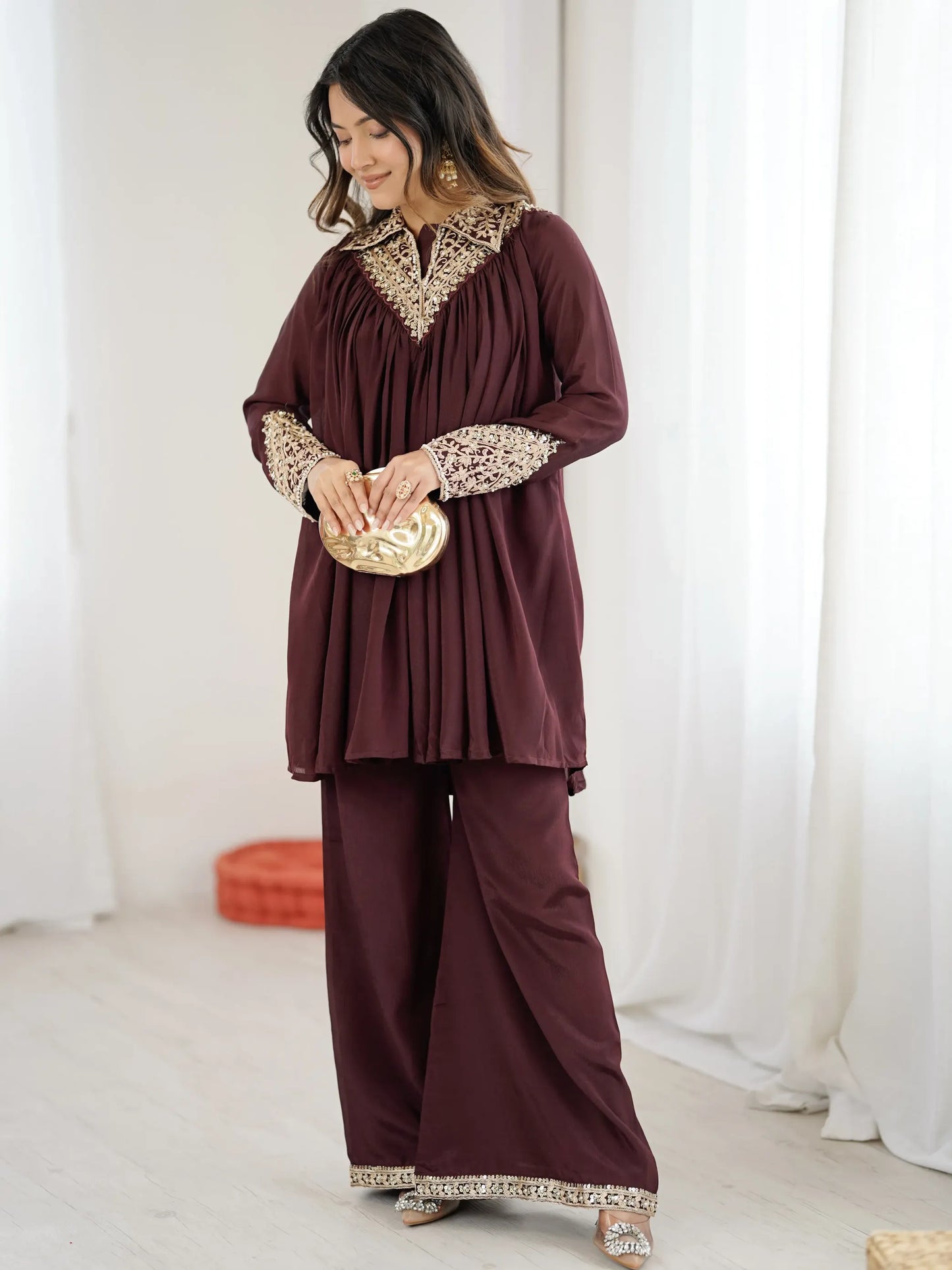 Woman wearing a burgundy outfit with gold embroidery against a white curtain background