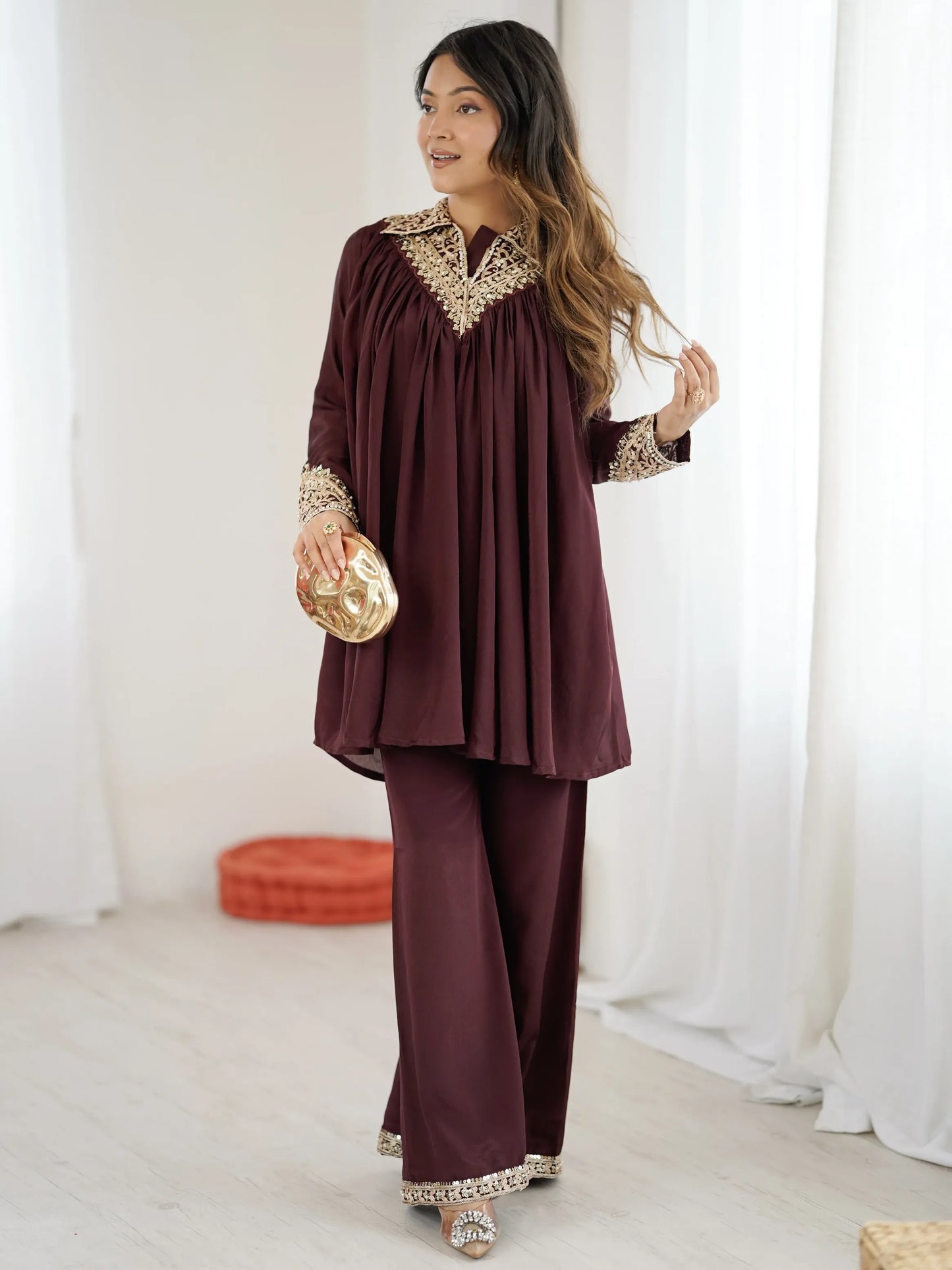 Woman wearing a burgundy outfit with gold embroidery holding a clutch against a white background