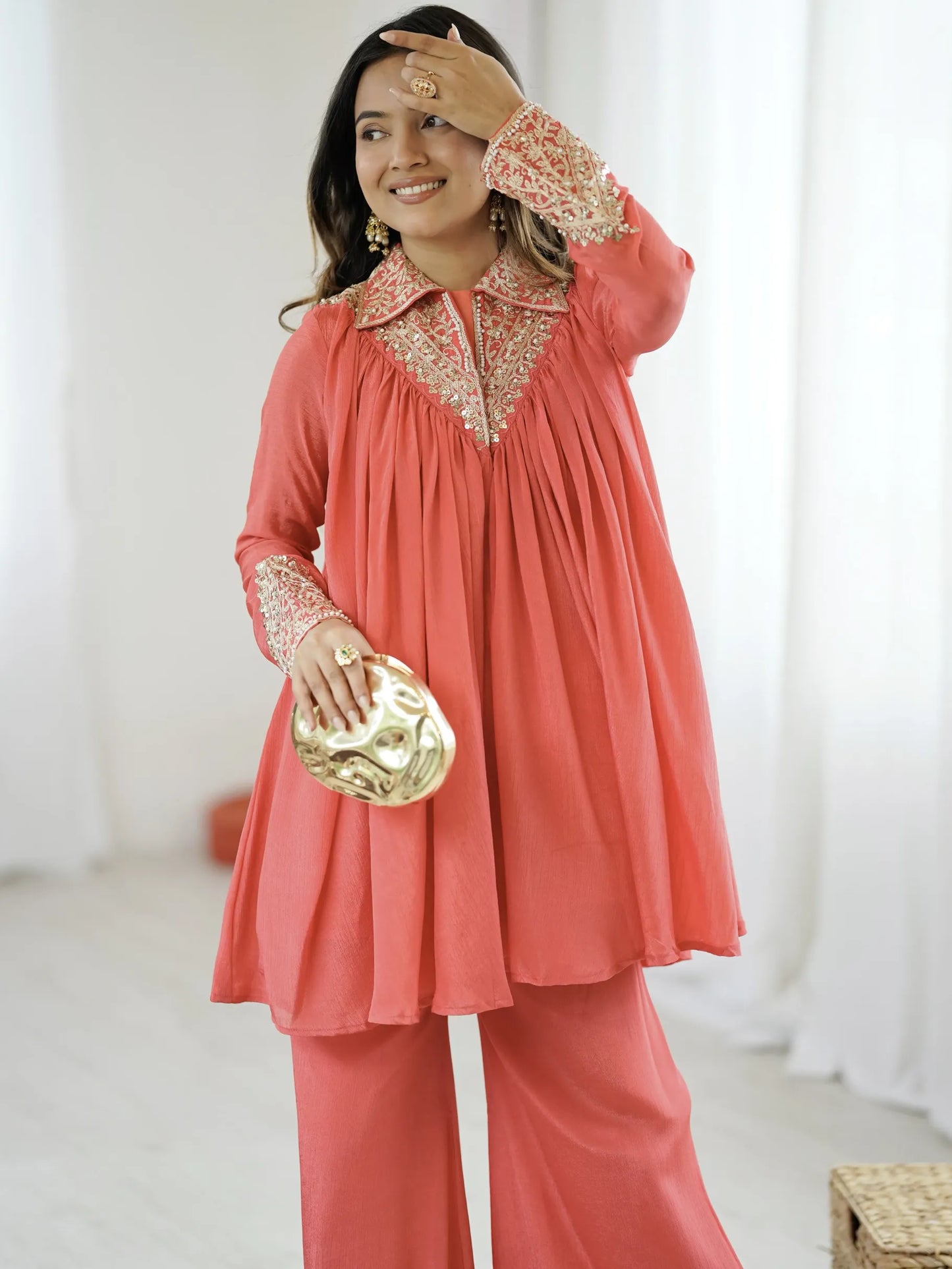 Woman wearing a coral outfit with intricate detailing, holding a gold clutch.