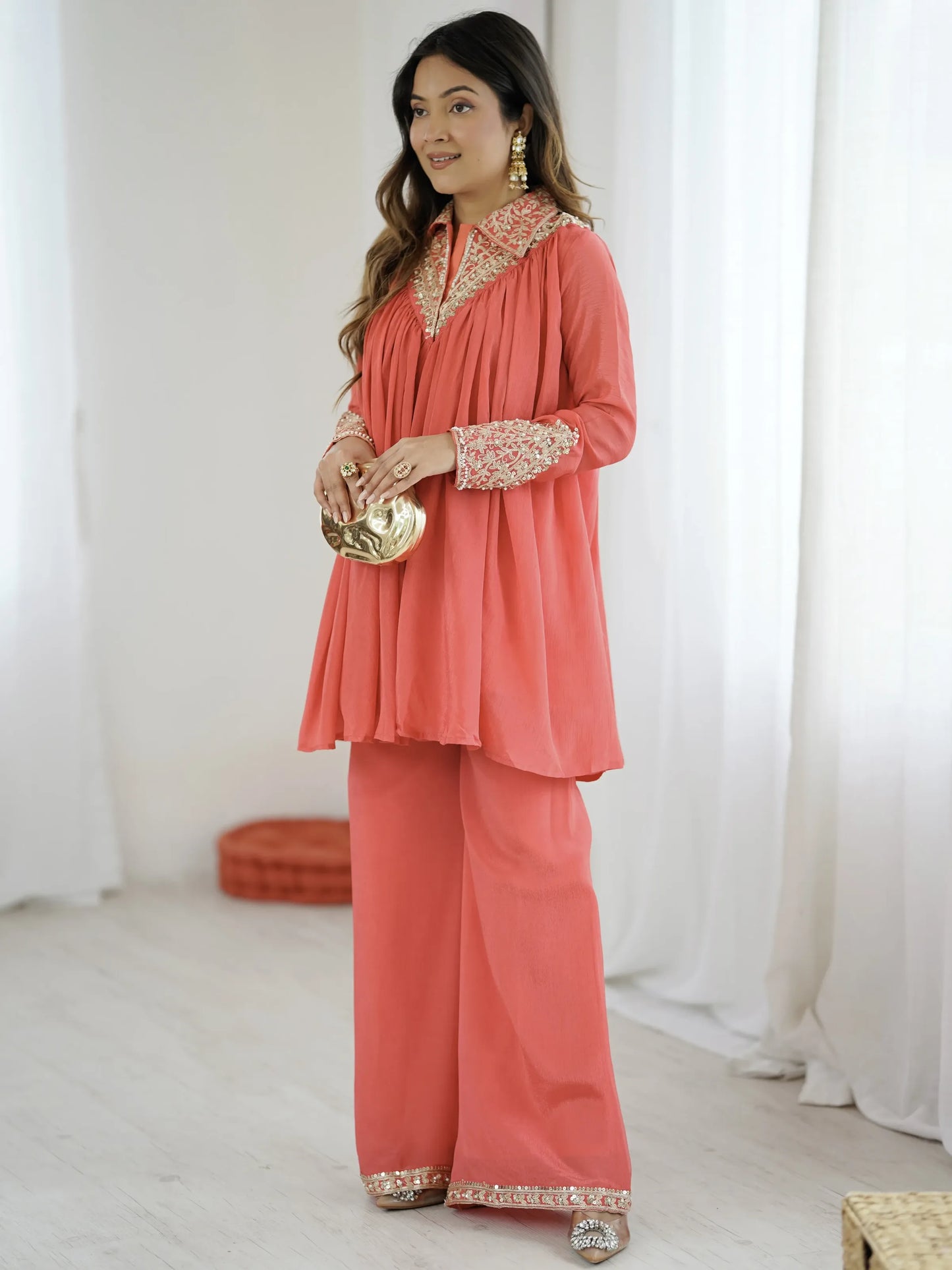 Woman wearing a coral traditional outfit with gold embroidery in a white room.