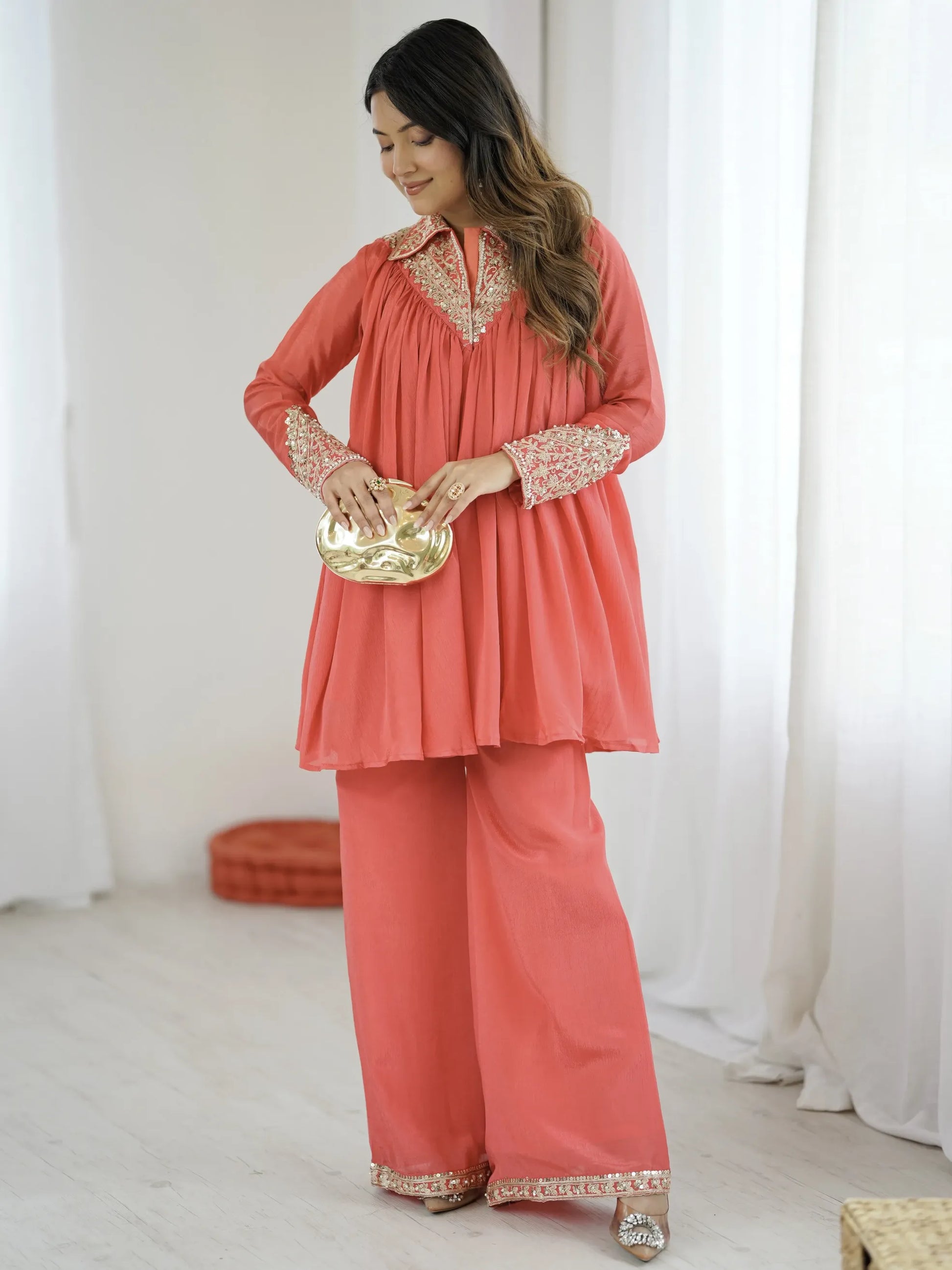Woman wearing a coral traditional outfit with white embroidery, holding a gold plate against a white curtain background.