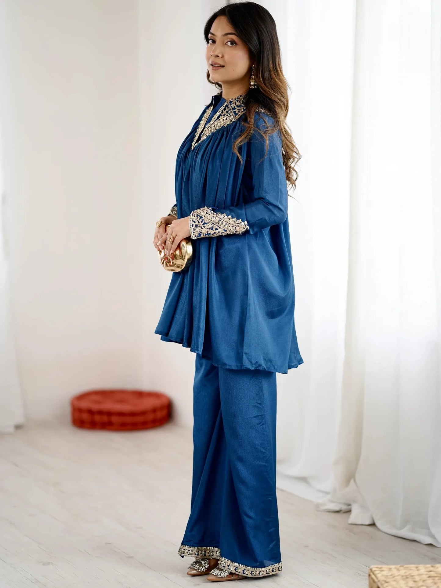 Woman in a blue traditional outfit standing in a room with white curtains and a red cushion.