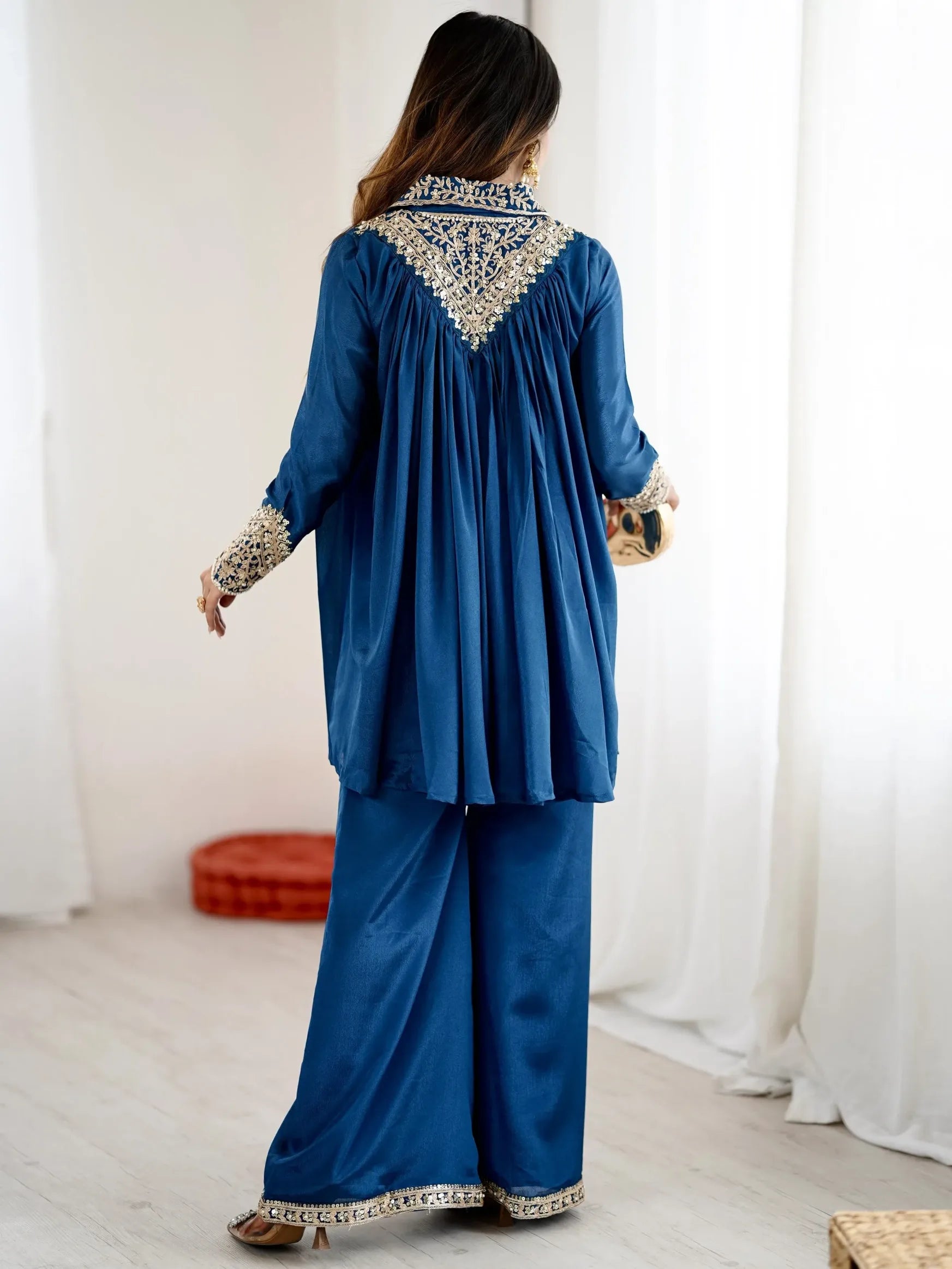 Woman wearing a blue traditional outfit with intricate patterns in a room with white curtains.