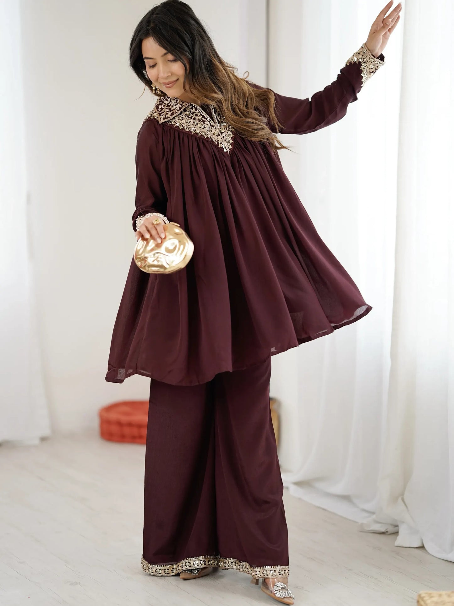 Woman in a burgundy traditional outfit with gold trim in a white room.