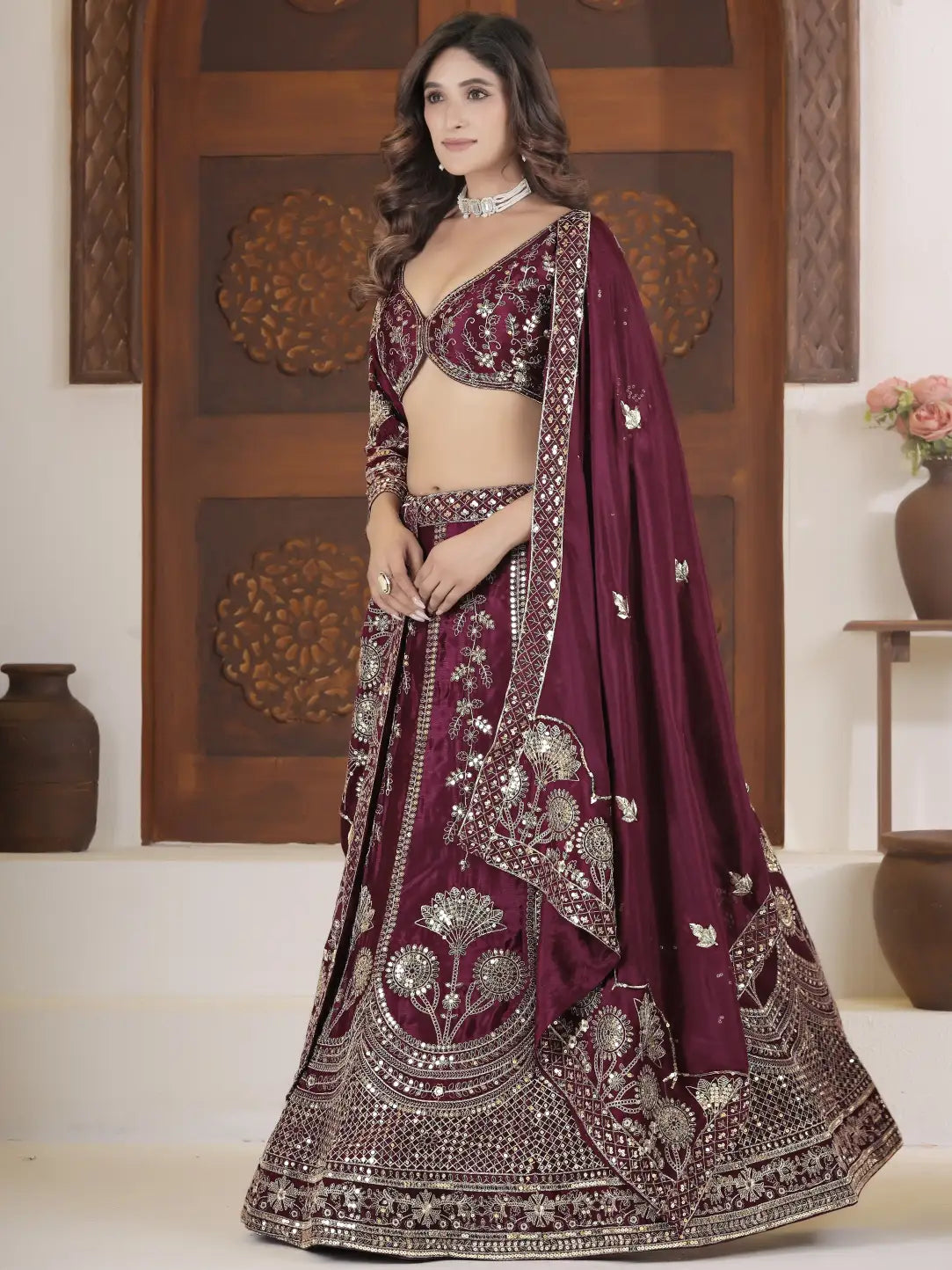 Woman in a maroon embroidered traditional outfit with silver details against a decorative wooden door.