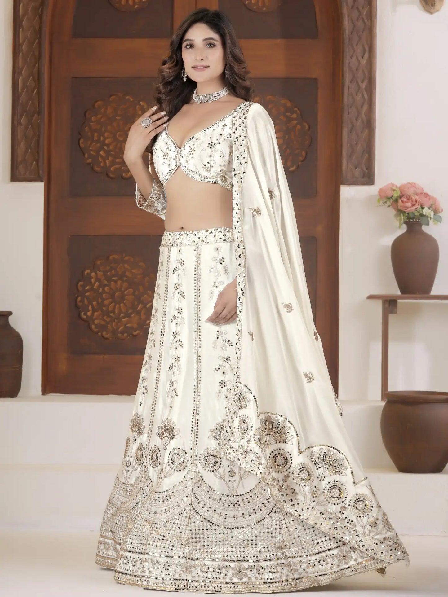 Woman in a white embroidered lehenga with a wooden panel and vase in the background