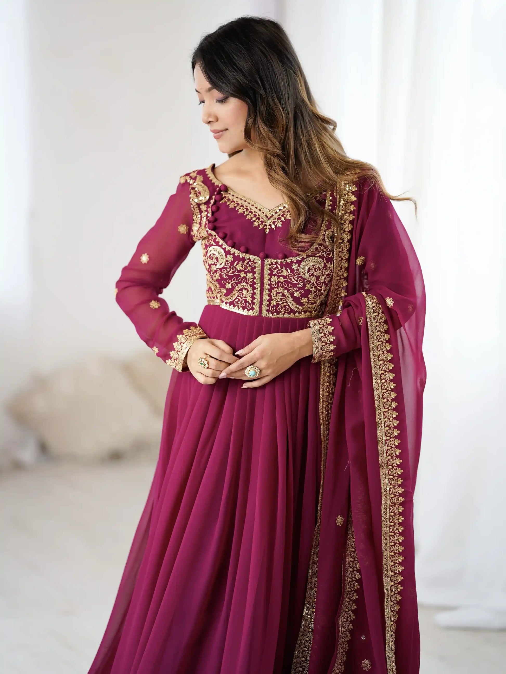 Woman wearing a magenta traditional outfit with gold embroidery in a minimal indoor setting.