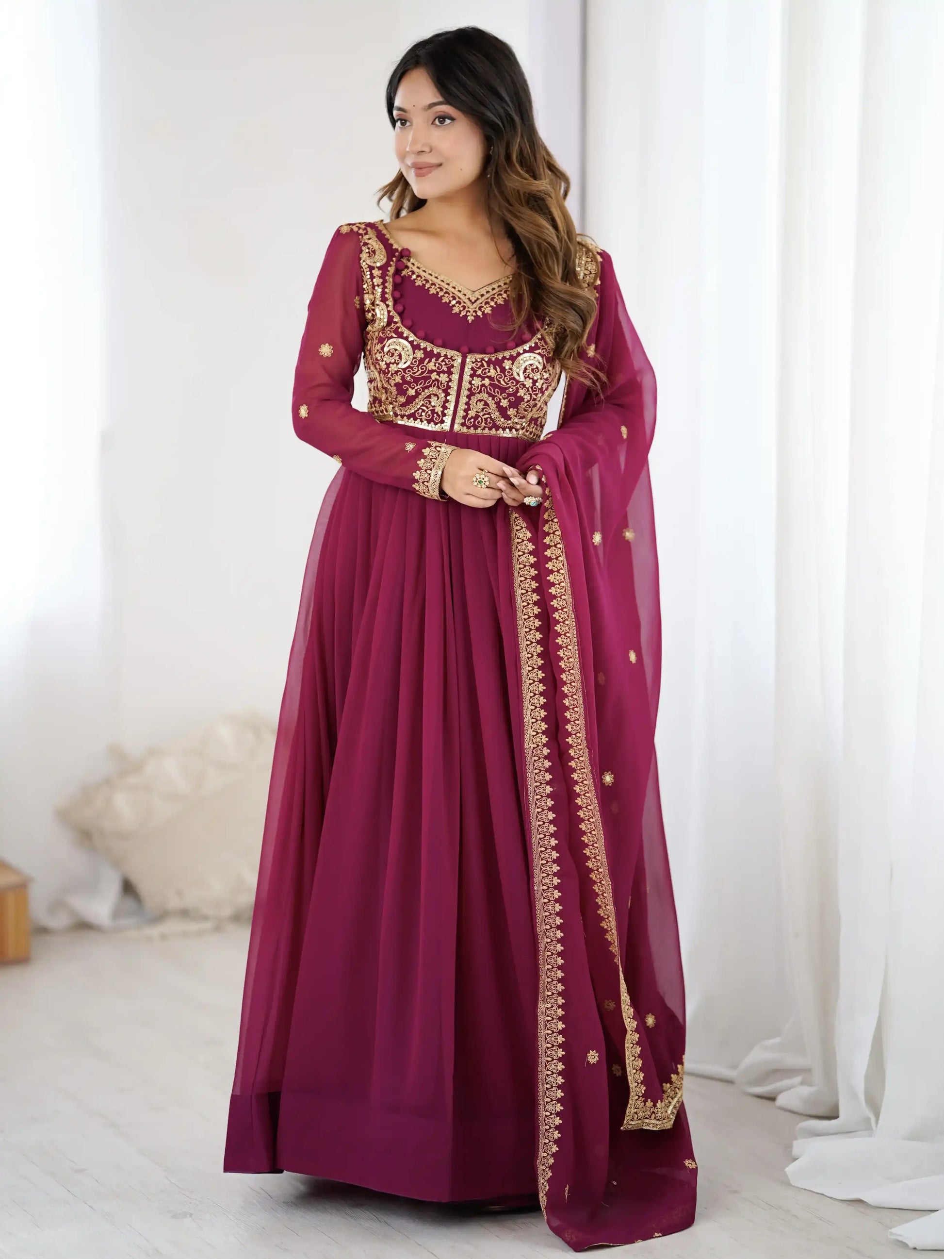 Woman wearing a maroon traditional outfit with gold embroidery in a white room.
