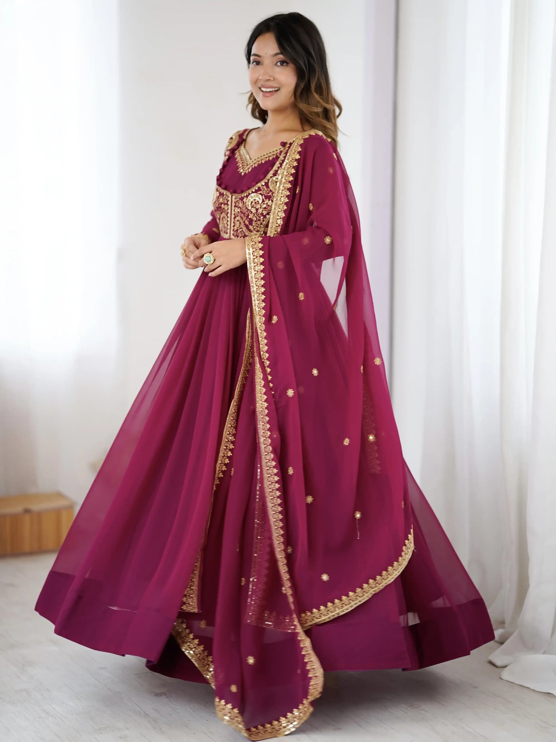 Woman wearing a maroon traditional outfit with gold embroidery against a white curtain background