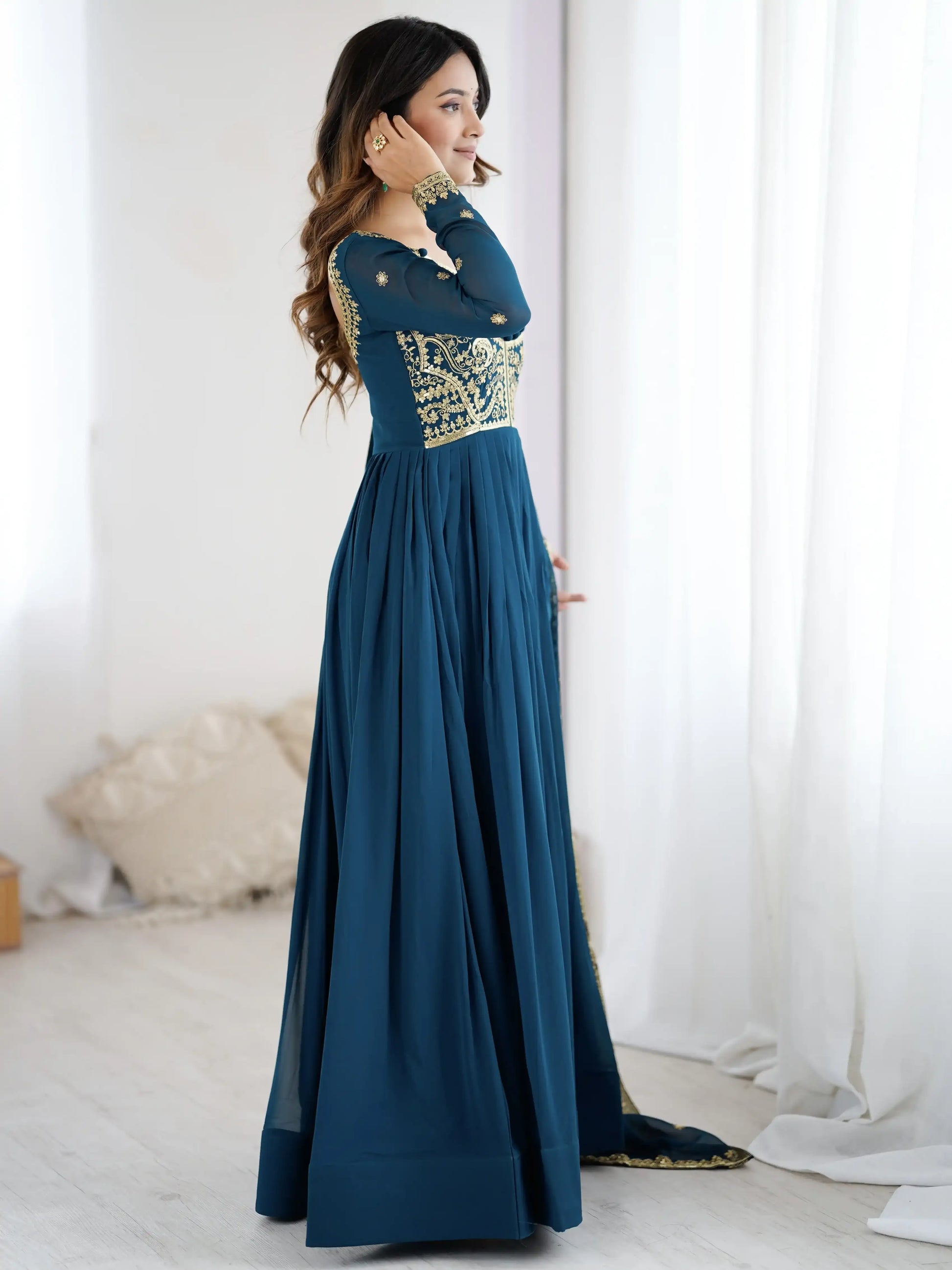 Woman wearing a long teal dress with gold embroidery in a room with white curtains.