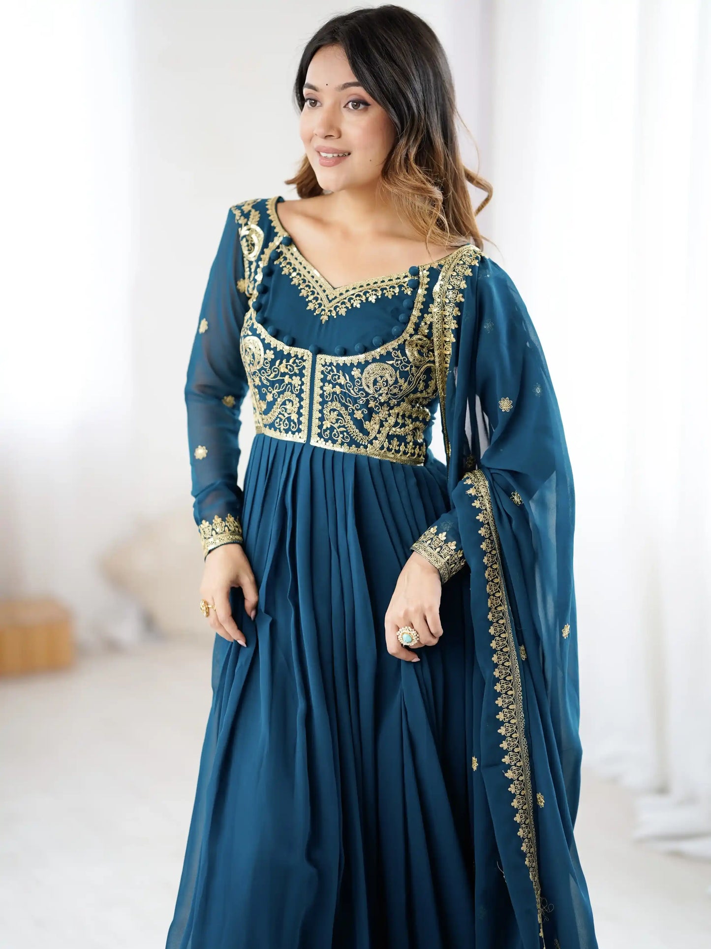 Woman wearing a teal traditional outfit with gold embroidery in a bright room.