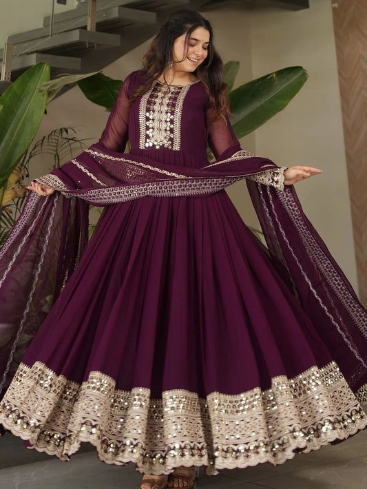 Woman wearing a purple traditional outfit with intricate designs, standing indoors with plants in the background.