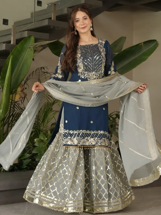Woman wearing a traditional blue and silver outfit with a white dupatta, standing indoors with plants in the background.