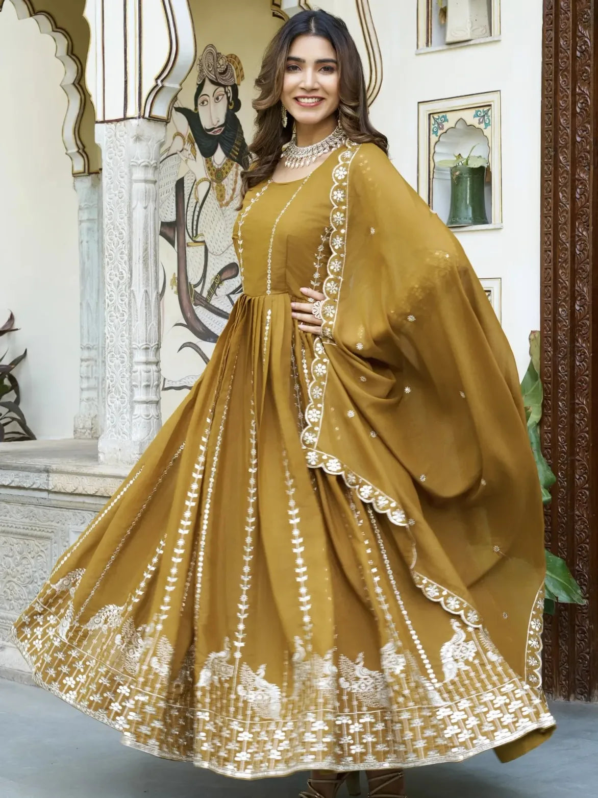 Woman in a mustard yellow traditional outfit with white embroidery standing in an ornate indoor setting.
