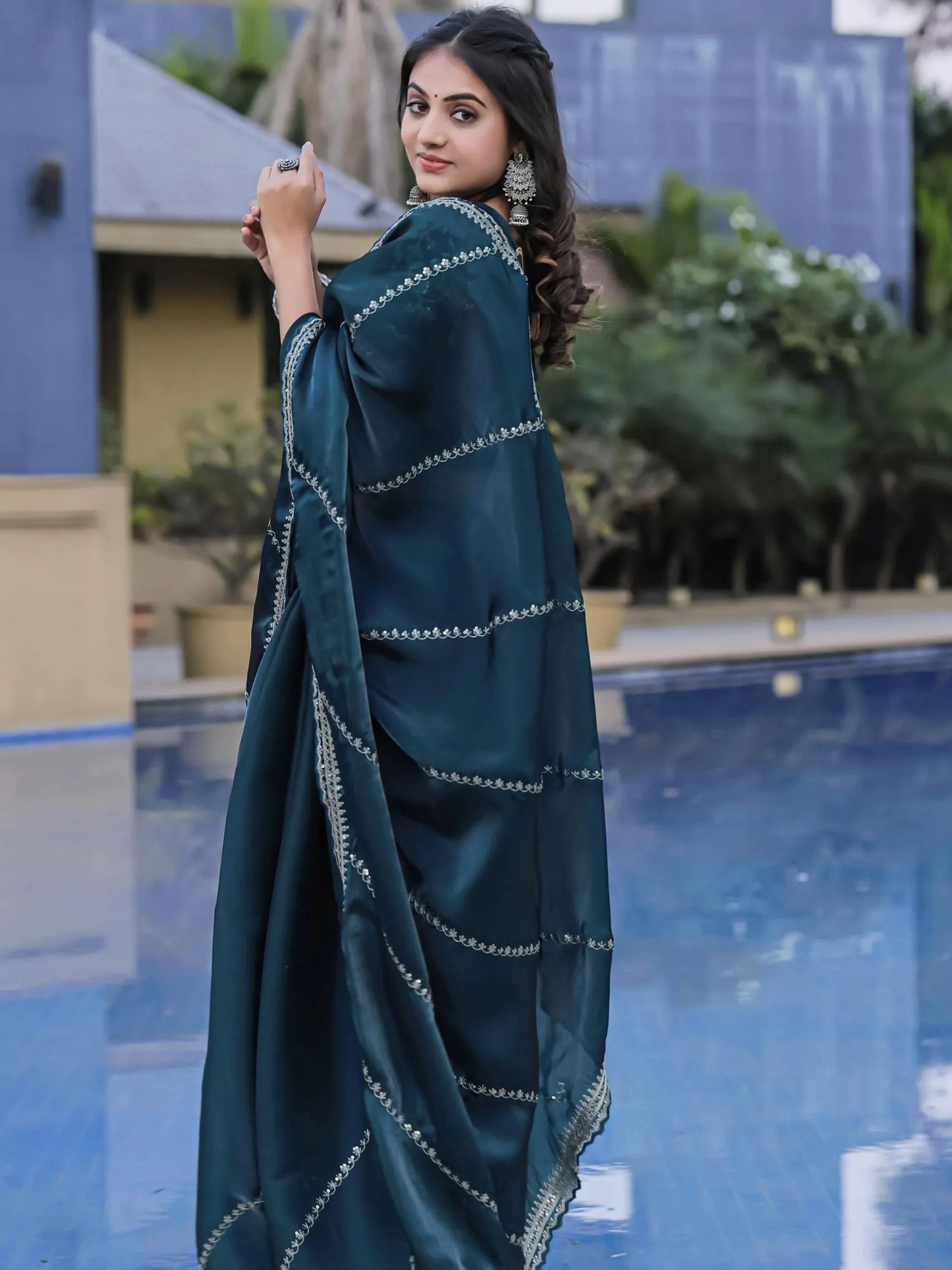Woman in a teal saree with white patterns standing by a pool.