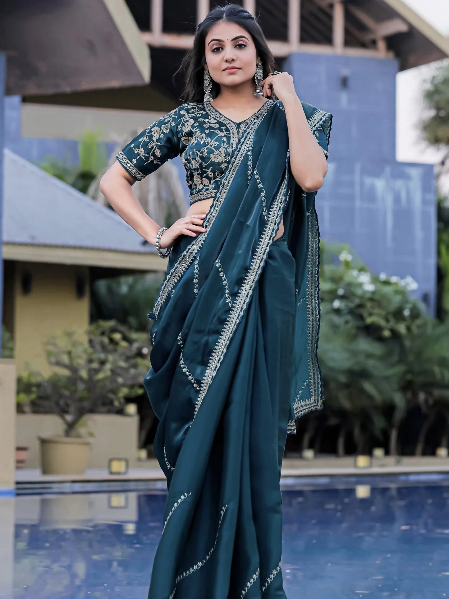 Woman in a teal saree with a matching blouse standing by a pool.