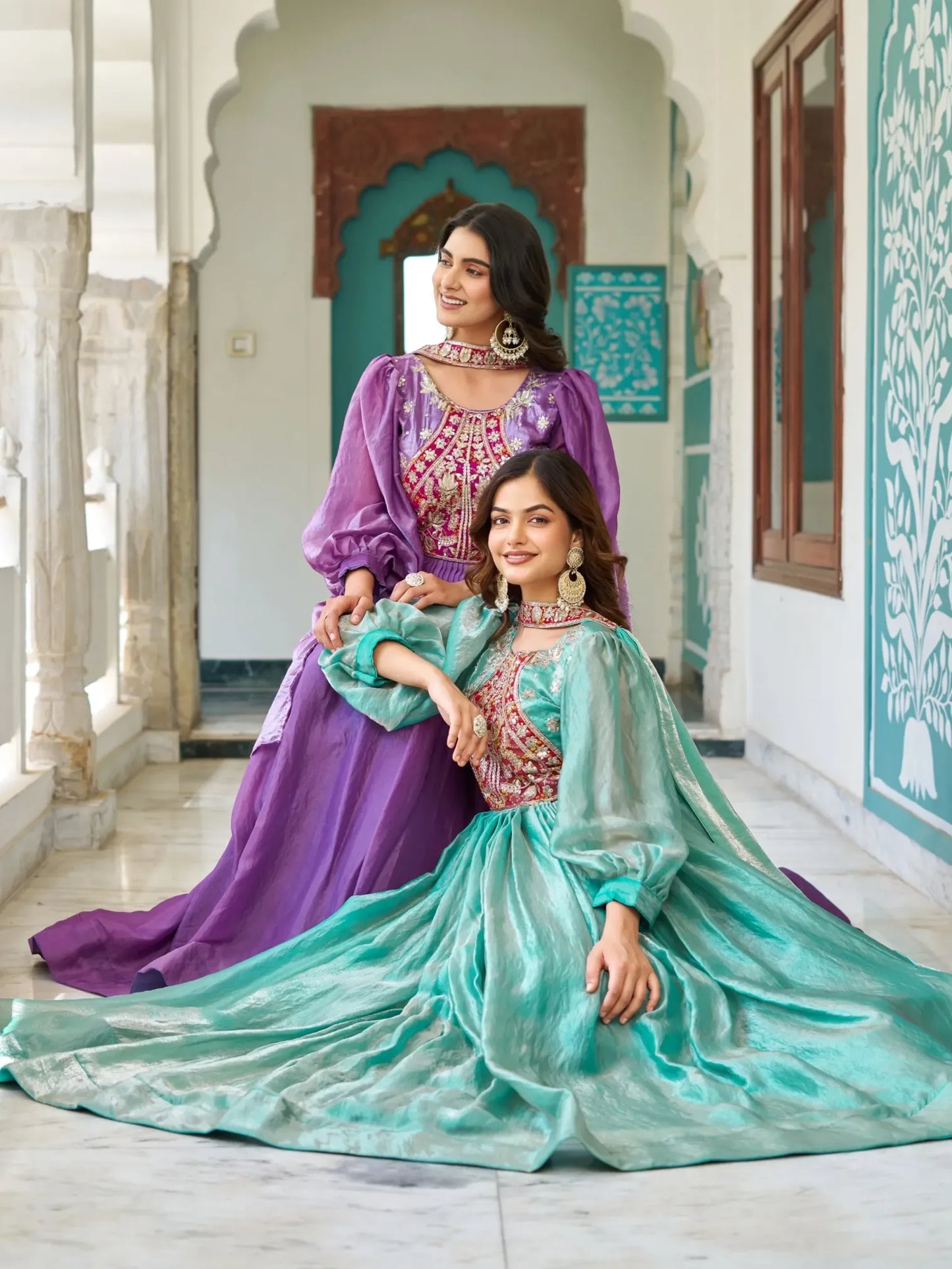 Two women in traditional outfits sitting in a decorative interior setting.