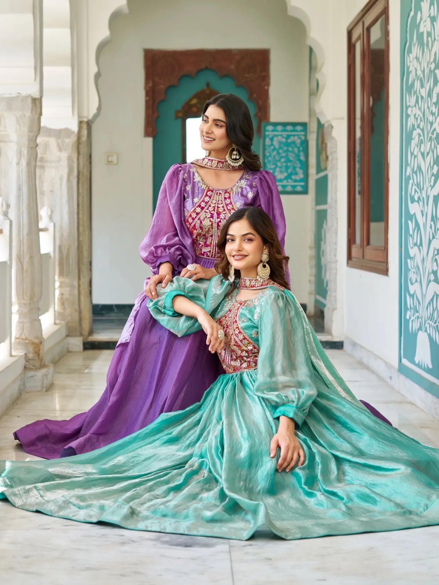 Two women in traditional outfits sitting in a decorative interior setting.
