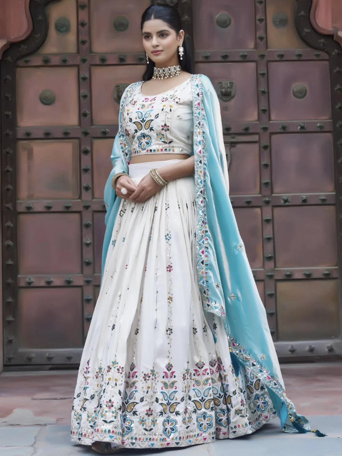 Woman in a traditional outfit with floral patterns standing in front of a metallic door.