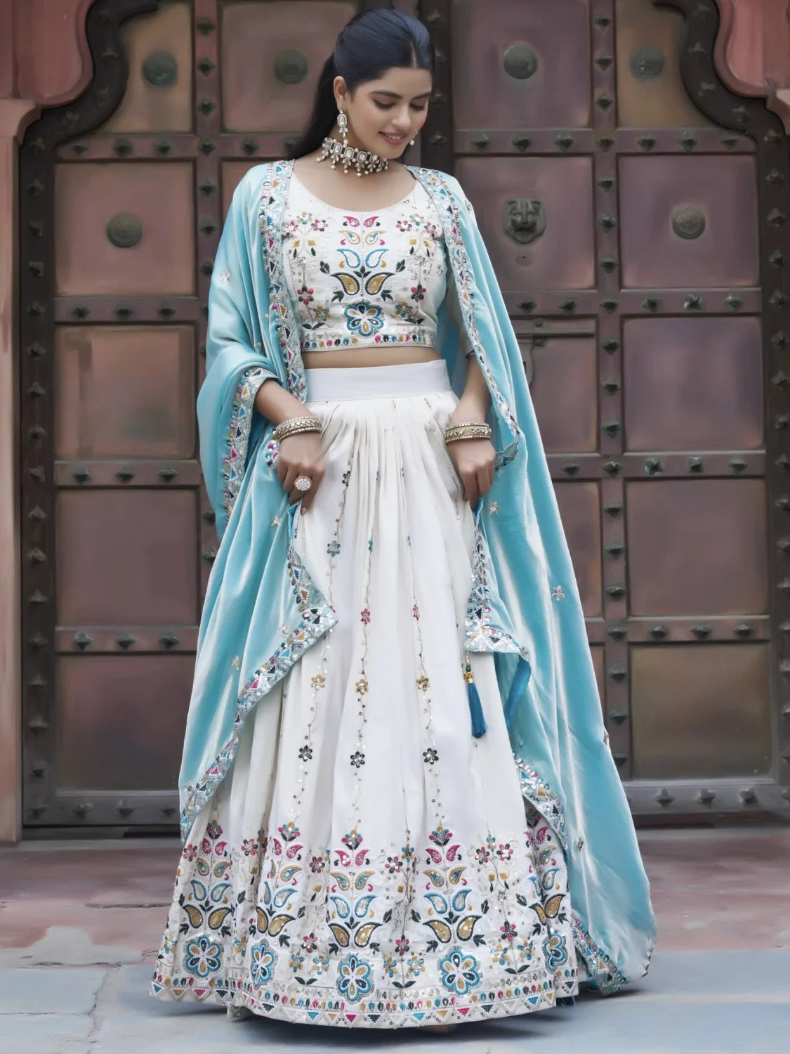 Woman in traditional embroidered outfit with light blue dupatta against a metallic door background